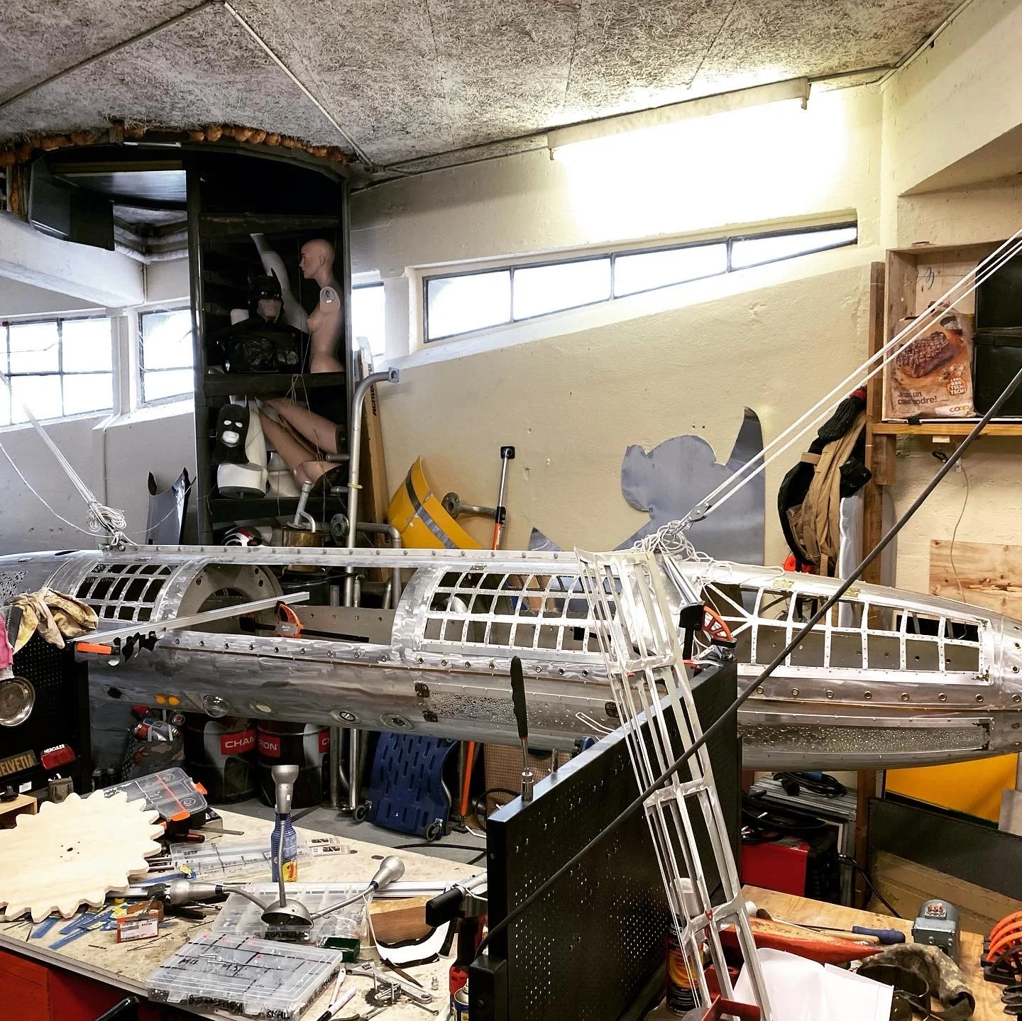 A cluttered workspace with an unfinished metallic airplane model, tools, and mannequin parts in the background.