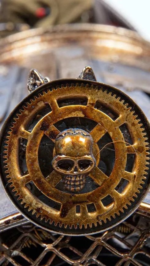 Close-up of a silver object with a gear and skull design, featuring small cat ears, placed on a wire mesh surface with blurred background.