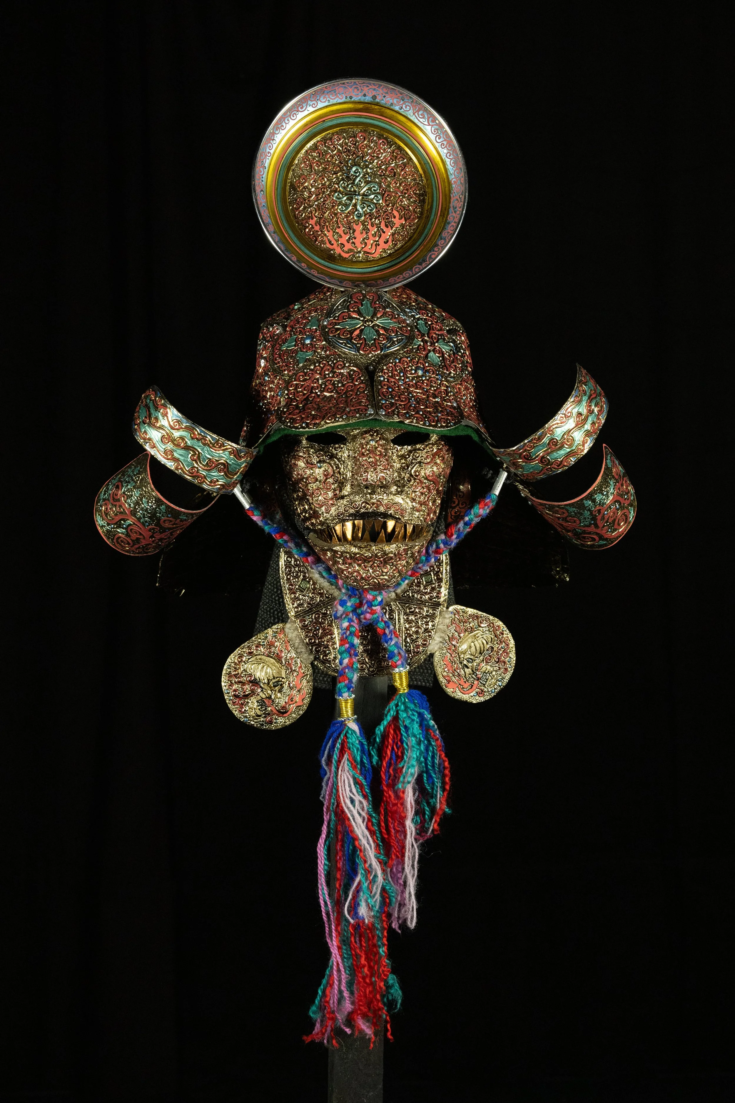 A traditional ornate mask with intricate designs, featuring a fierce face with gold teeth and an elaborate headdress with embroidery, beads, and adornments. The mask is displayed against a black background.