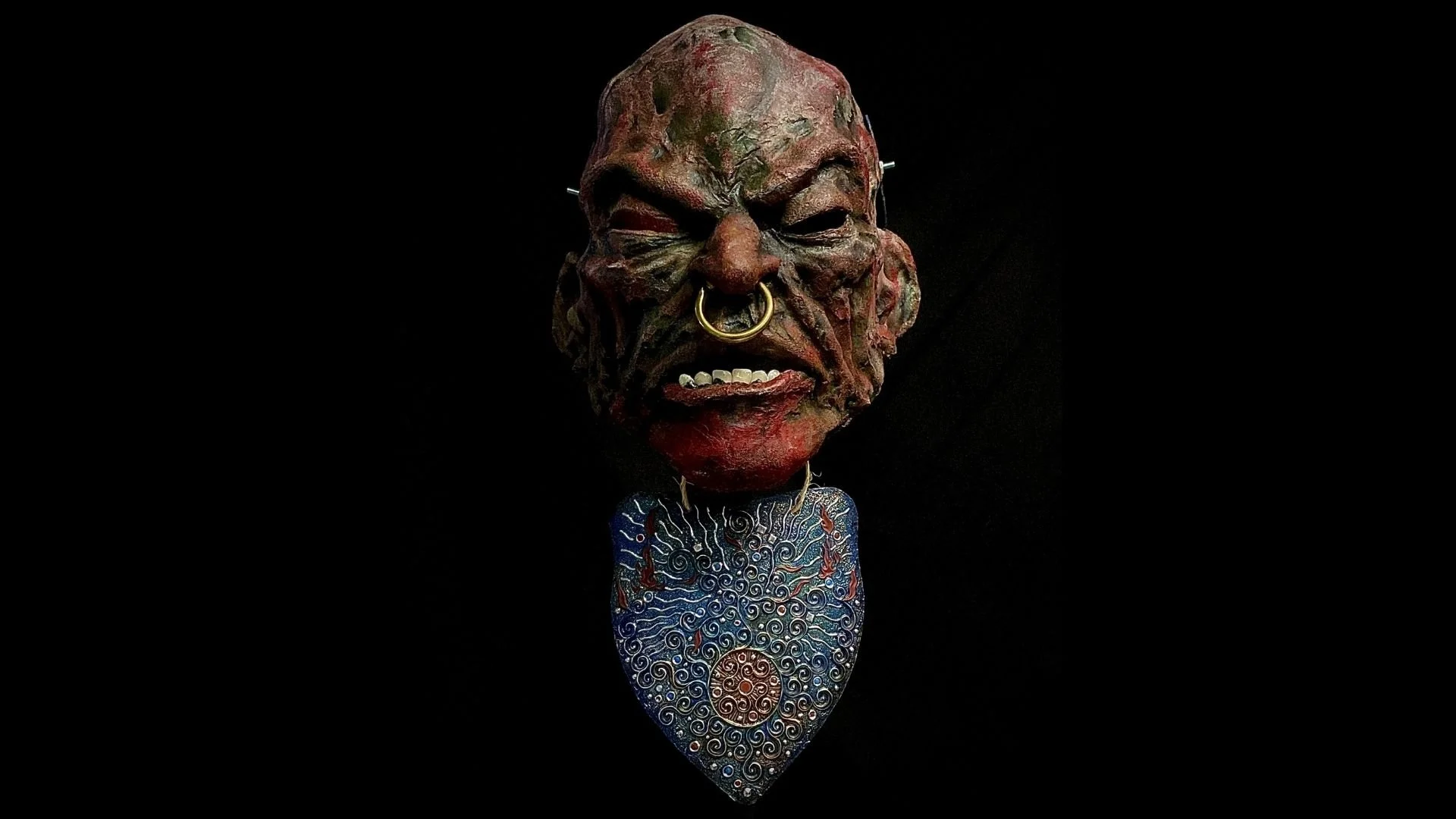A tribal mask with exaggerated facial features, dark skin tone, and piercings, mounted on a black background, with colorful necklaces hanging below.