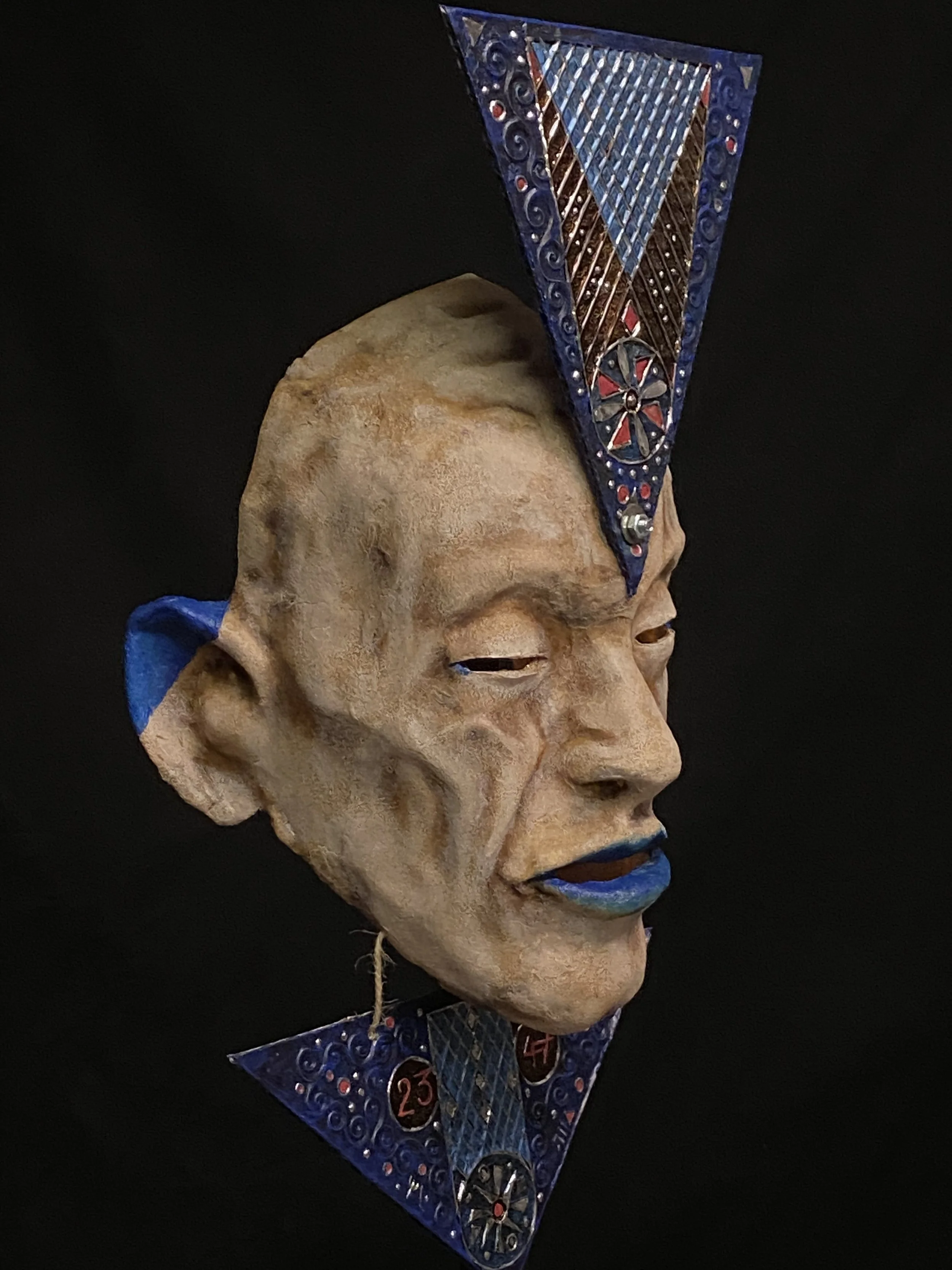 Mixed media art piece featuring a human face sculpture with closed eyes and blue lips, adorned with metal and colorful geometric structures on top and below the head, against a black background.