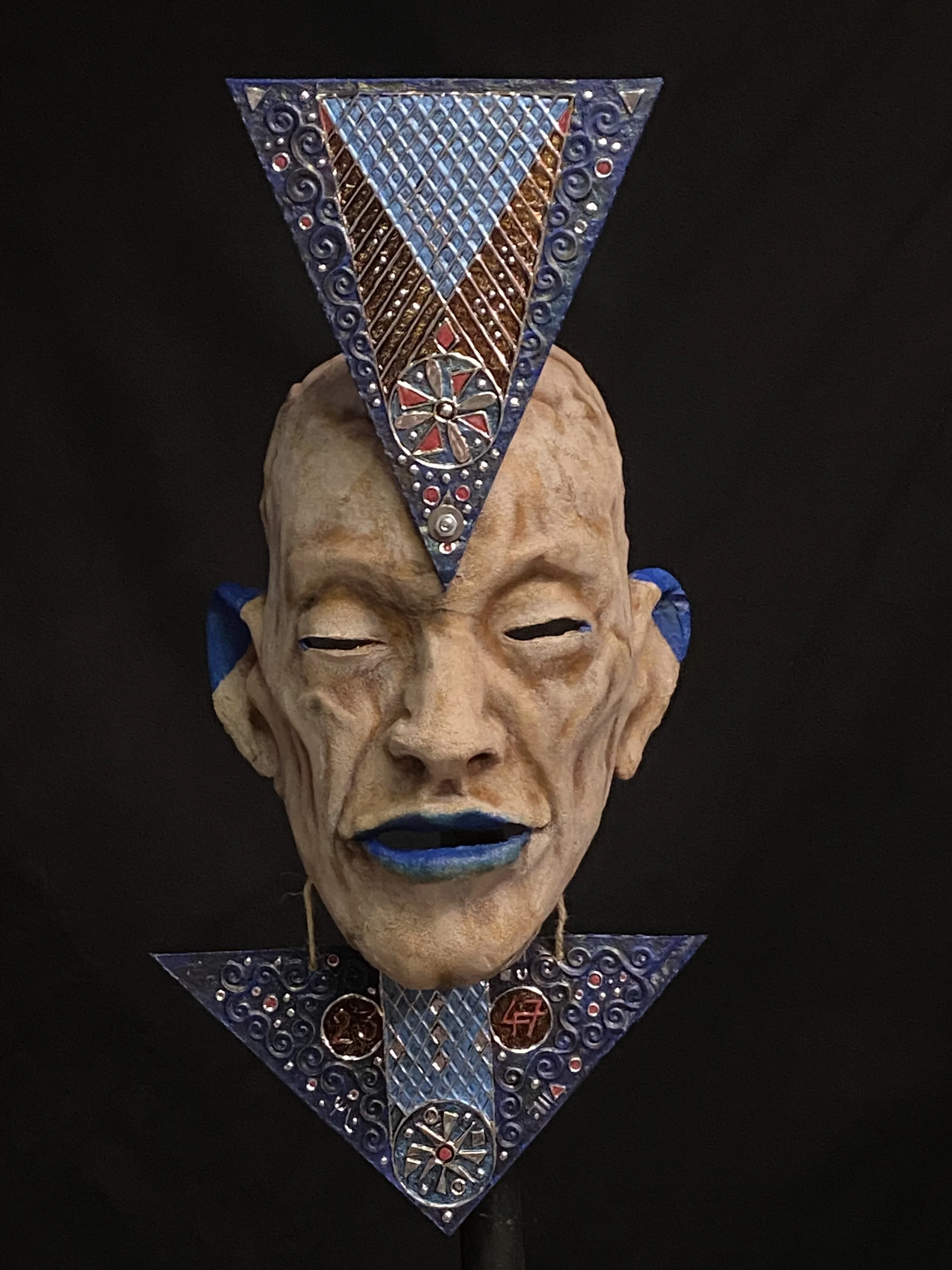 A mask with a human face, featuring a beige complexion, blue lips, closed eyes, and blue accents on the ears, decorated with ornate geometric designs and red and silver embellishments, set against a black background.