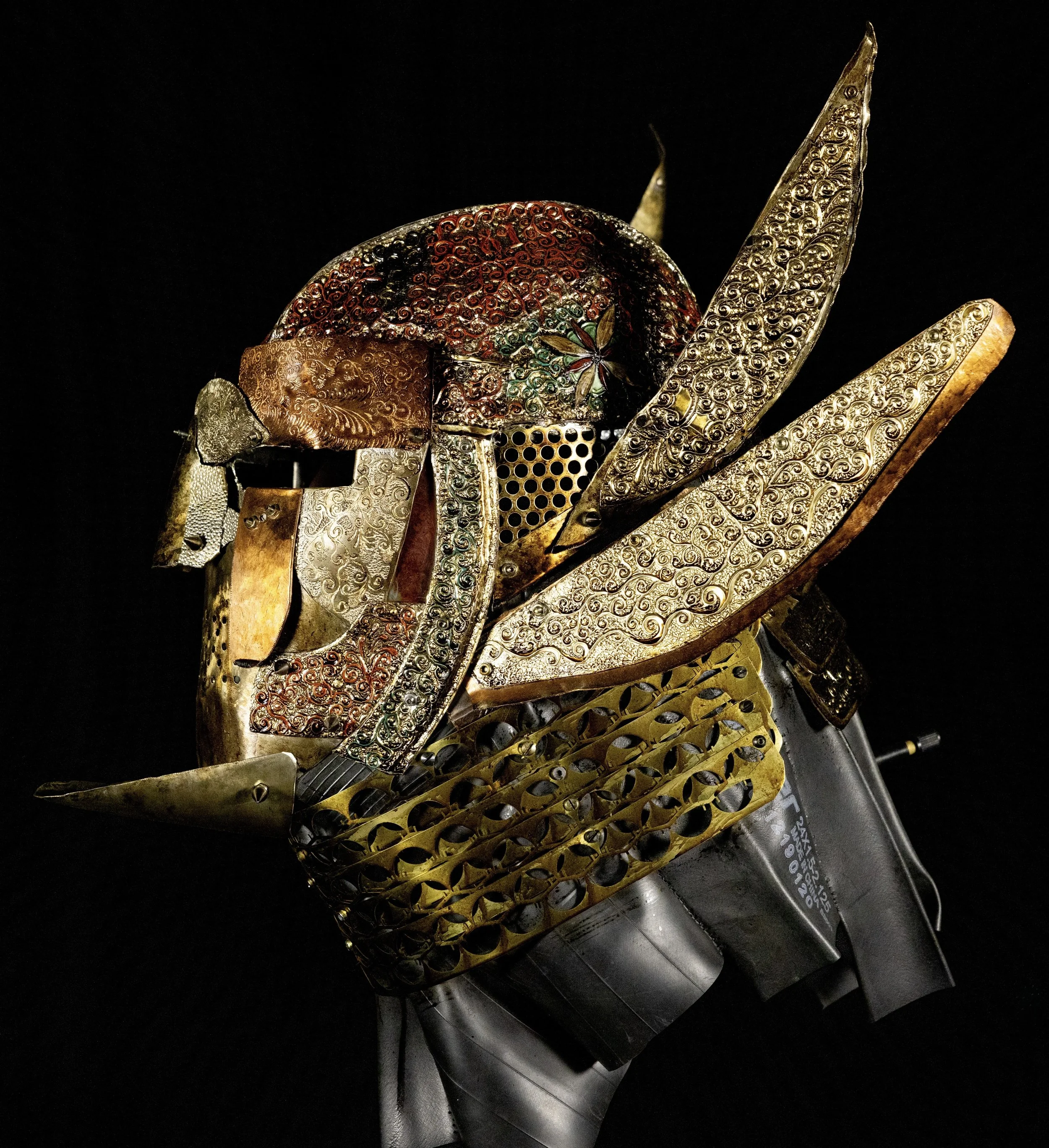 An artistic sculpture of a warlord helmet created from intricately designed metallic and gold elements, with a black background.