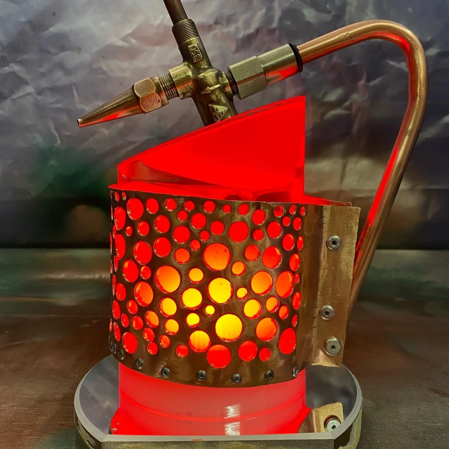 A scientific or industrial device with a perforated metal body, glowing red interior, and a brass pipe with a pointed tip and a gauge, set on a metallic base.