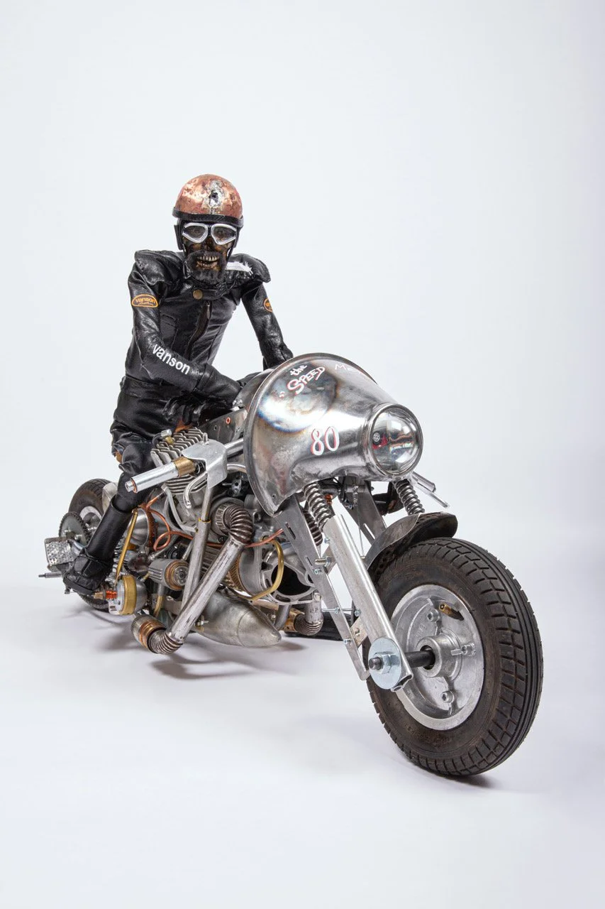A skeleton riding a vintage motorcycle with a large headlight, wearing a black leather jacket, gloves, goggles, and a shiny pink helmet, against a plain white background.