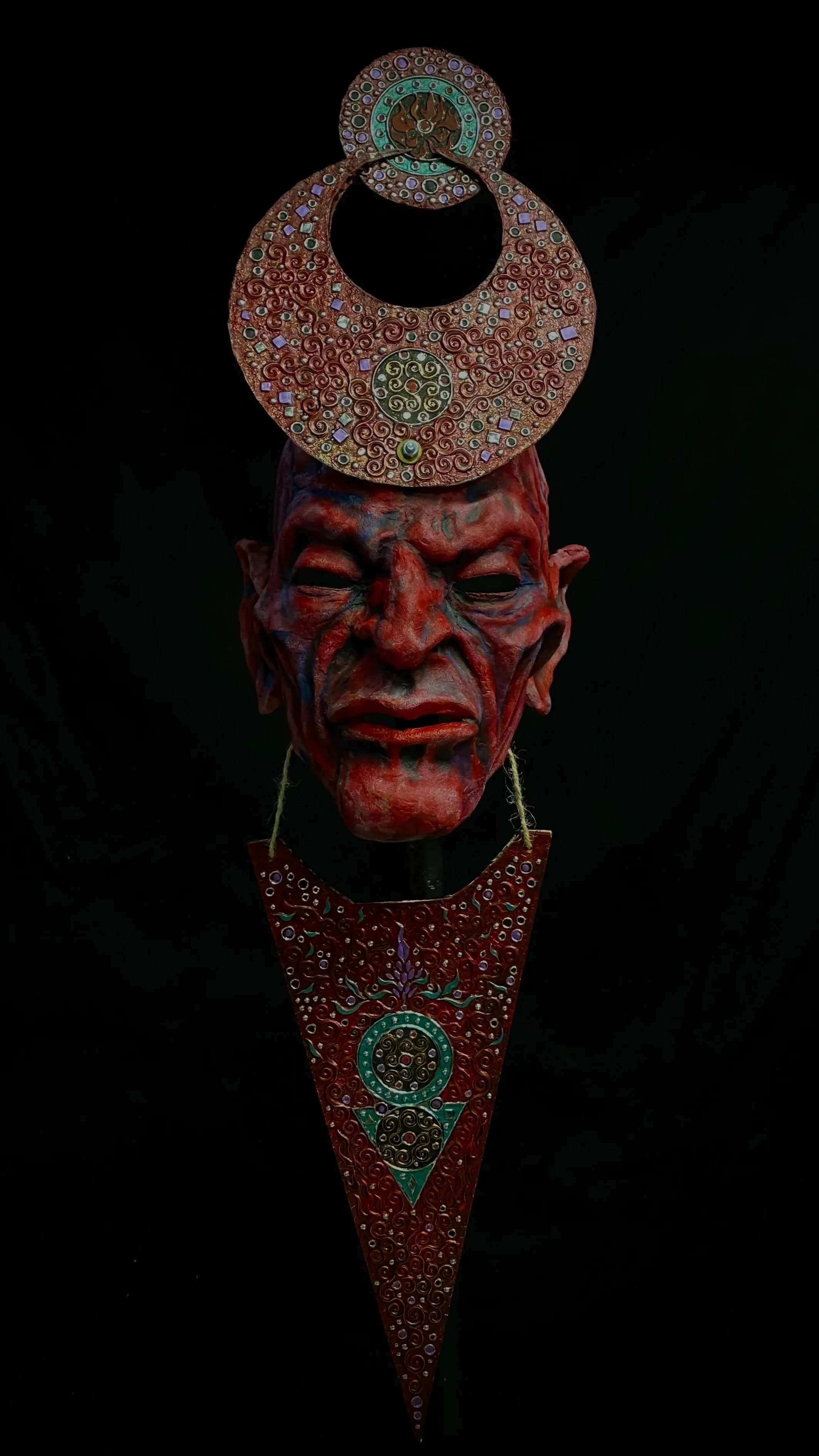 A mask of an elderly person's face with red and black paint and a headpiece with intricate circular designs, including a large circular ornament on top and another below, connected by a string.