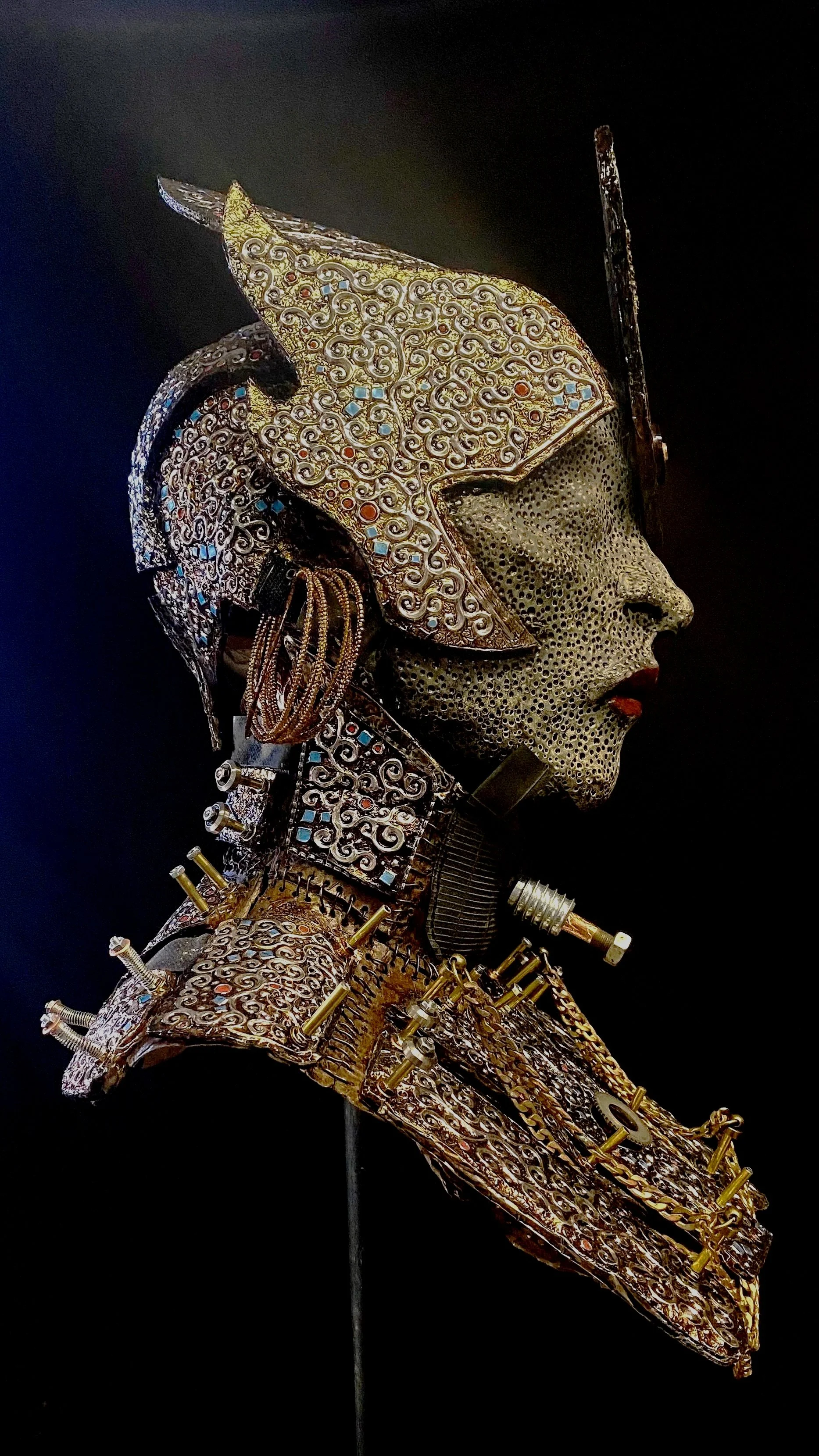 A detailed sculpture of a woman’s profile with intricate metalwork and jewelry, featuring a headdress and necklace with geometric and swirling patterns.