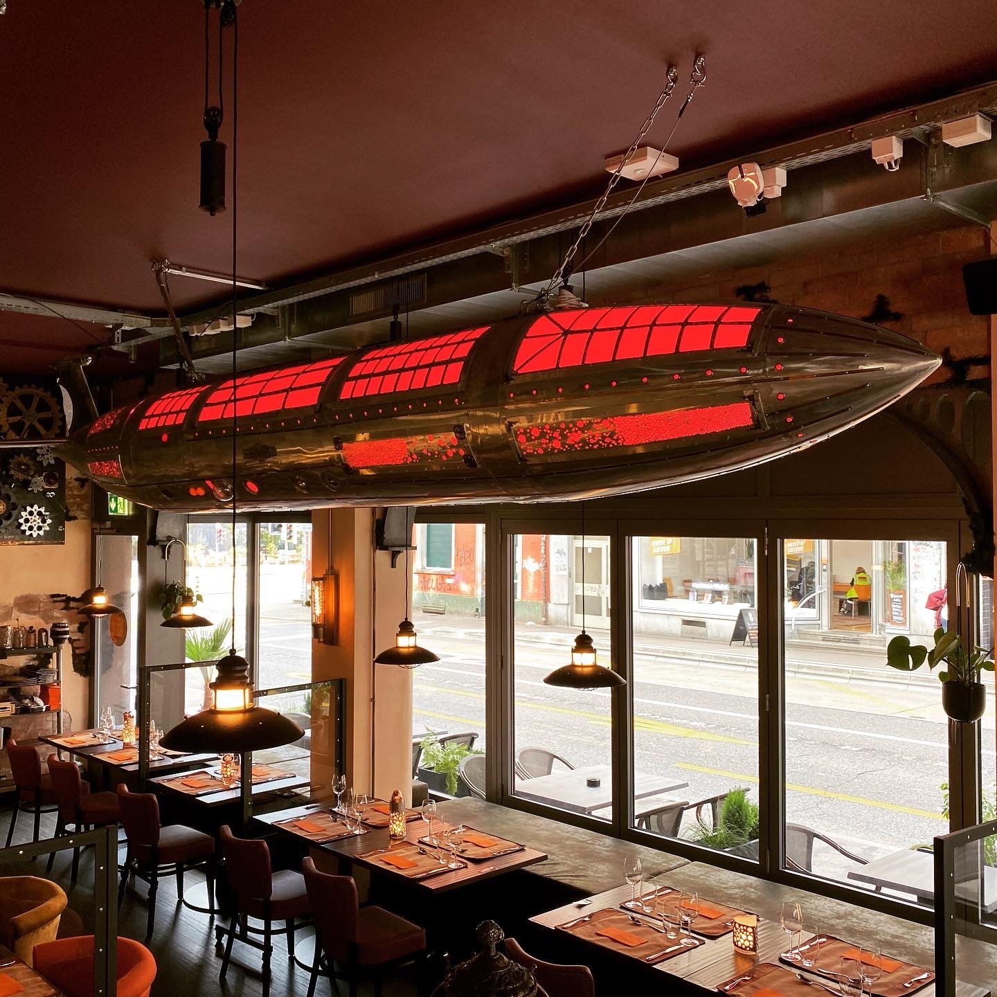 Interior of a restaurant with a unique rocket-shaped hanging light fixture, wooden tables set for dining, and large windows showing an outside street scene.