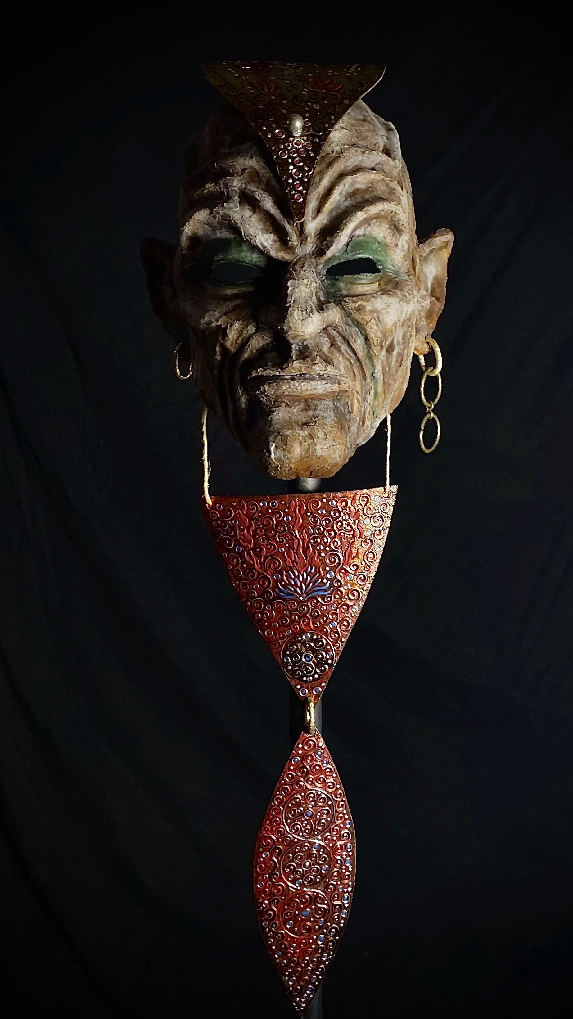 A traditional mask with carved wooden face, green eye shadow, earrings, and red decorative elements, set against a dark background.