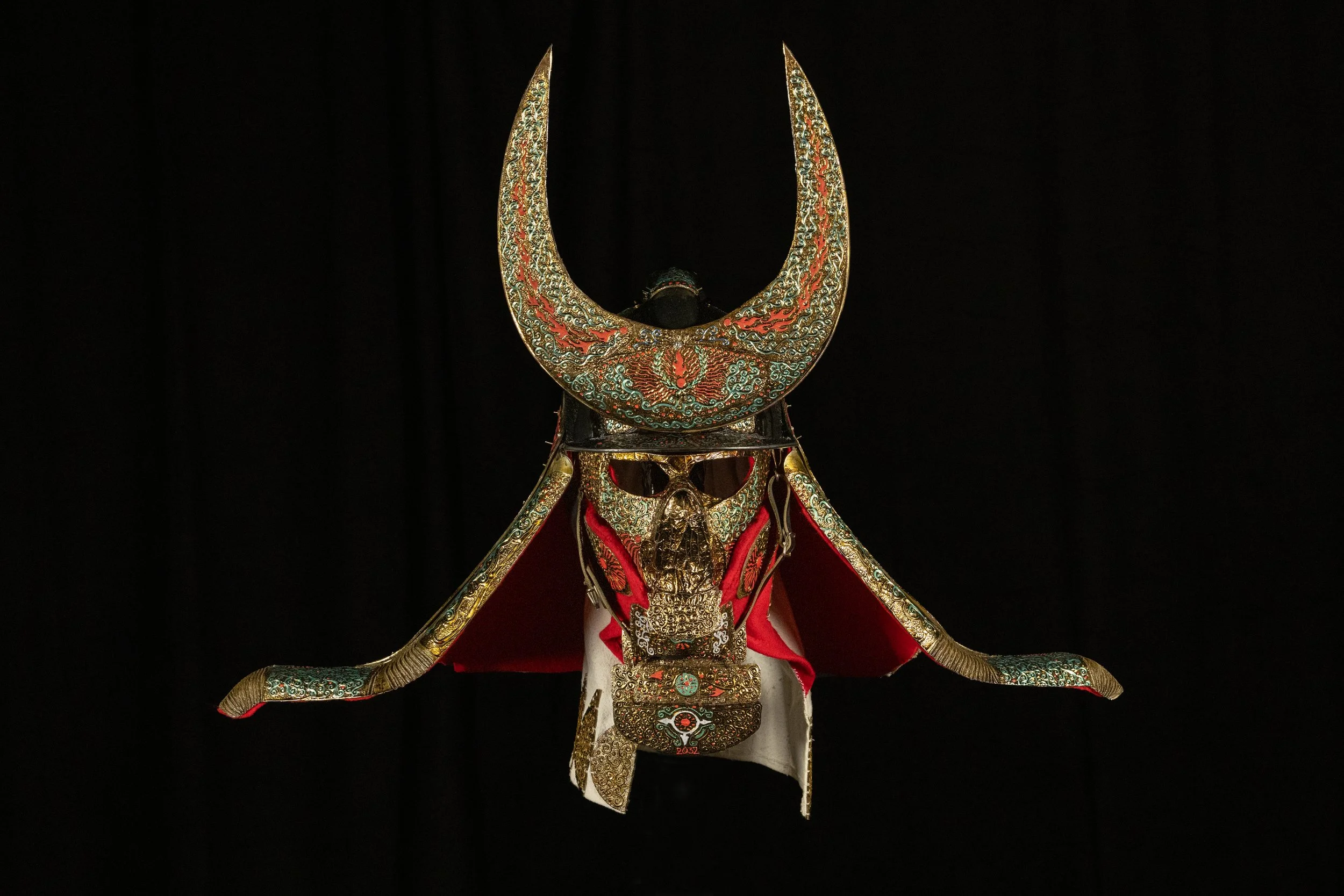 A decorative helmet with a large crescent-shaped ornament on top, adorned with intricate gold and red patterns, featuring a face mask with elaborate designs and a red cape behind it, set against a black background.