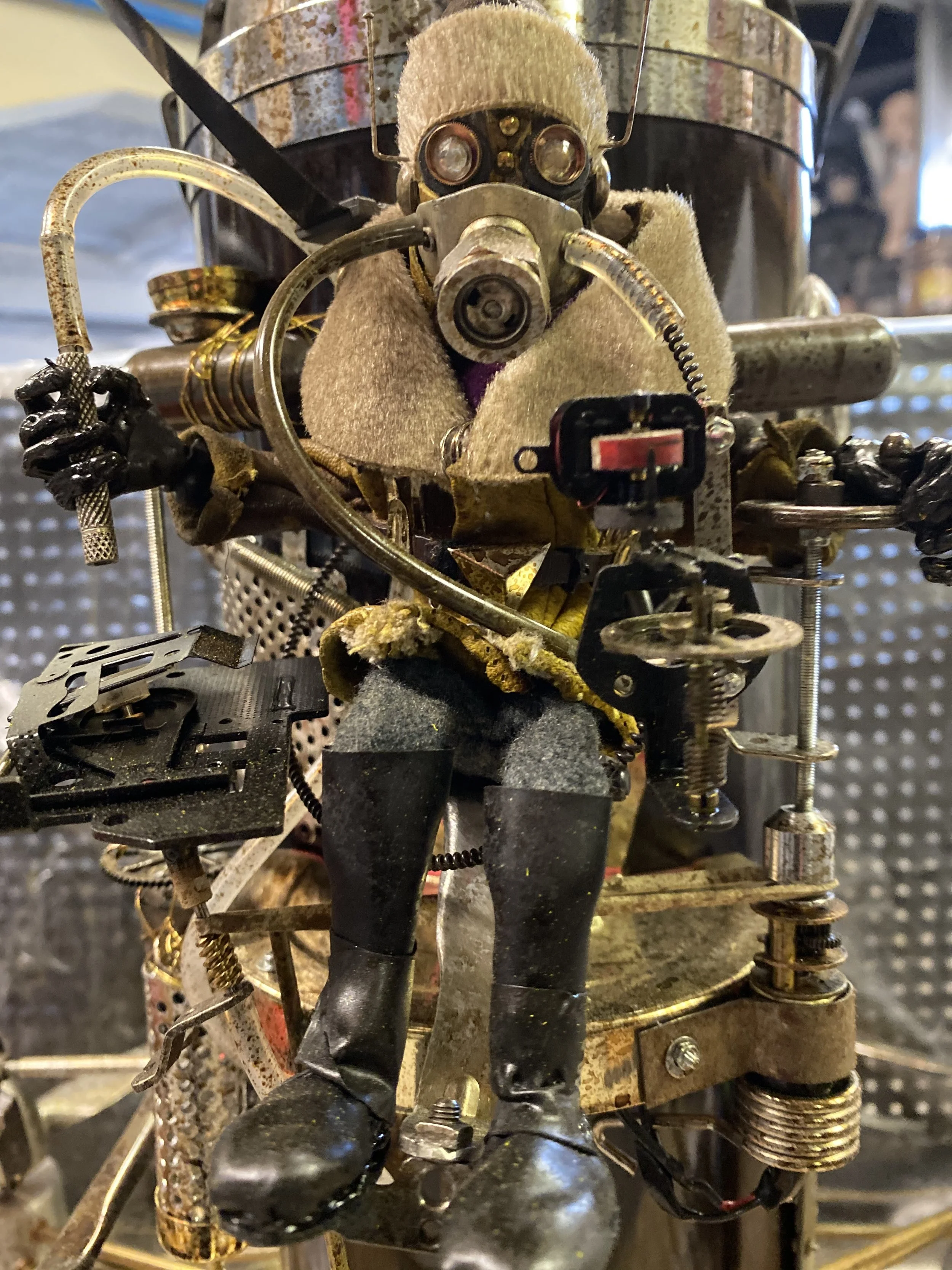 A robot made of metal parts with a small stuffed toy in a bee costume as its head, sitting in front of a black cylindrical object.