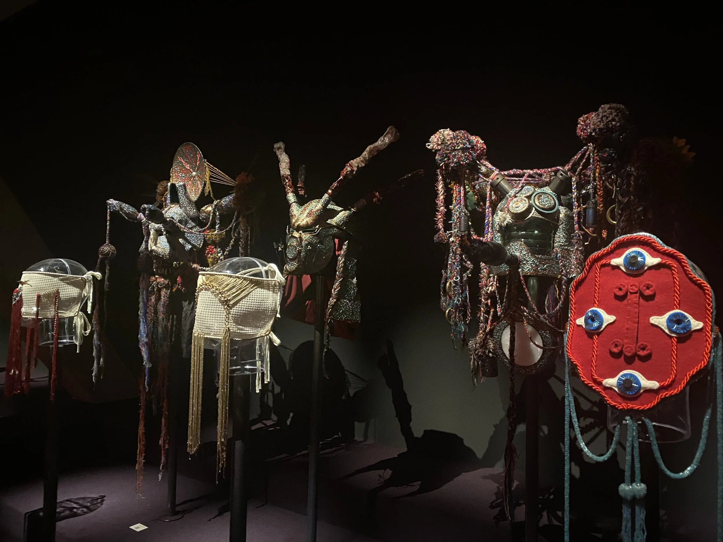 Display of elaborate tribal masks and headdresses adorned with beads, shells, and fabric in a museum setting.