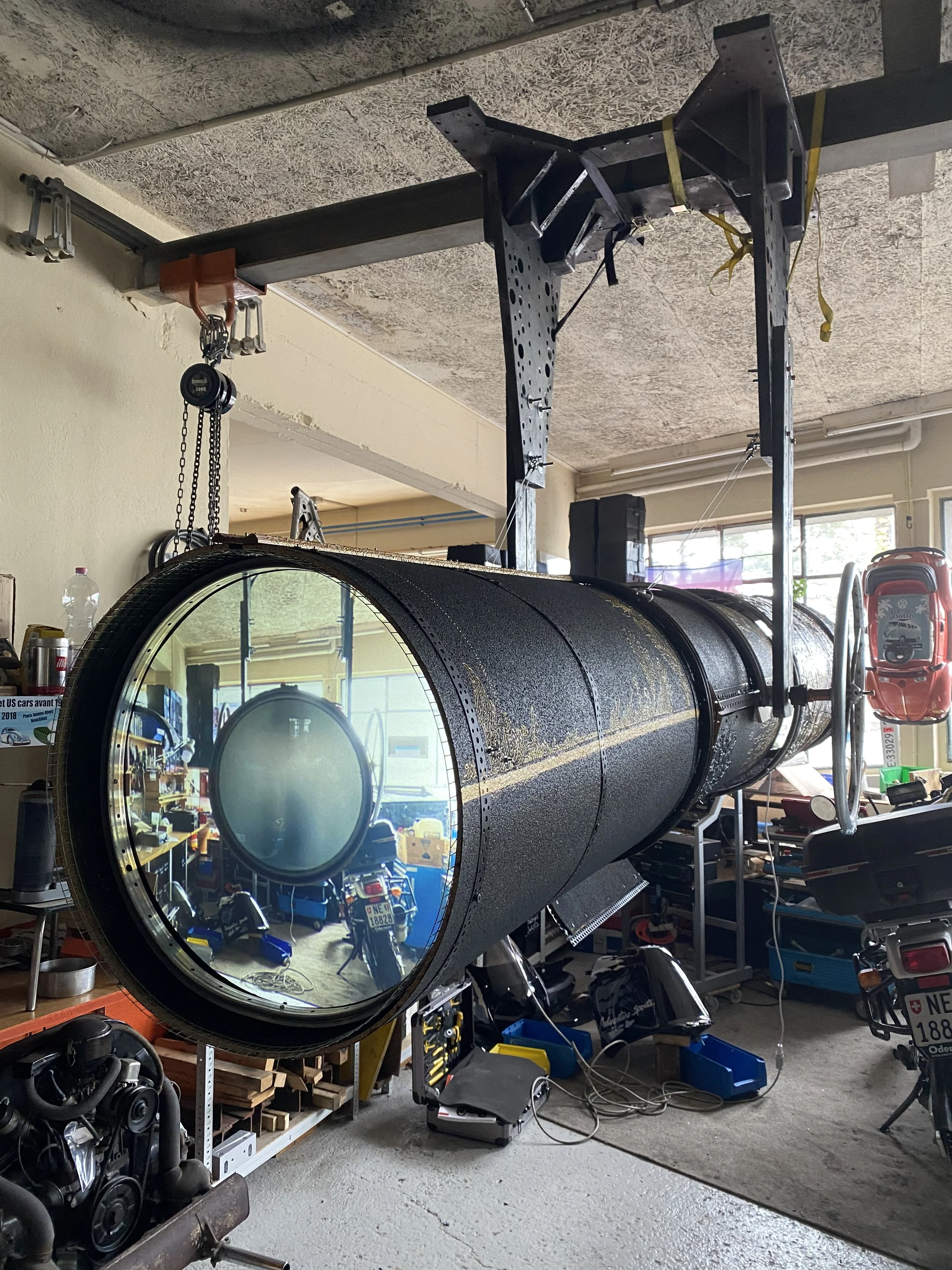 A large, black telescope mounted on a support structure in a garage or workshop, with various tools and equipment around.