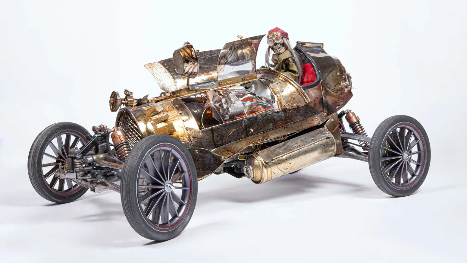 A vintage, steampunk-style racing car with a skeletal figure in the driver's seat, constructed from metal parts with exposed mechanical components, and four large wheels.