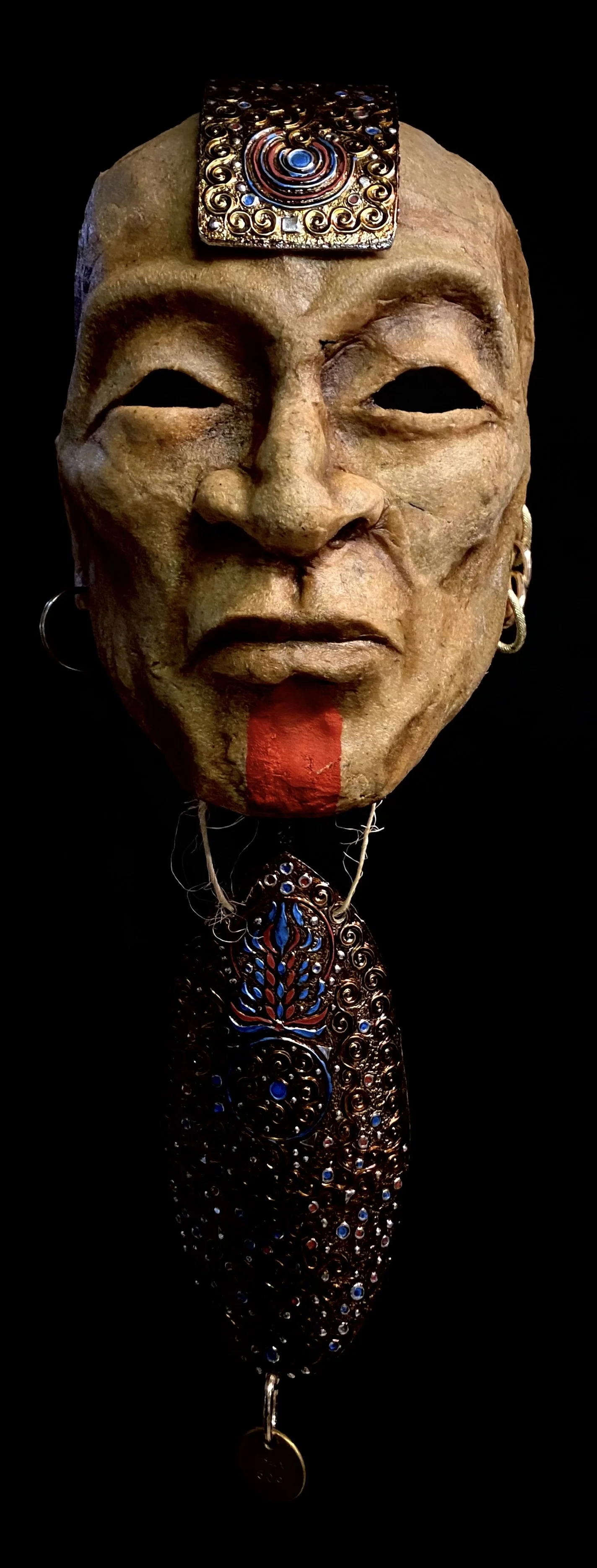 A tribal mask with a large face featuring pronounced features, adorned with ornate jewelry and decorations, including a detailed metallic element on the forehead and an elaborate beaded or metallic necklace.