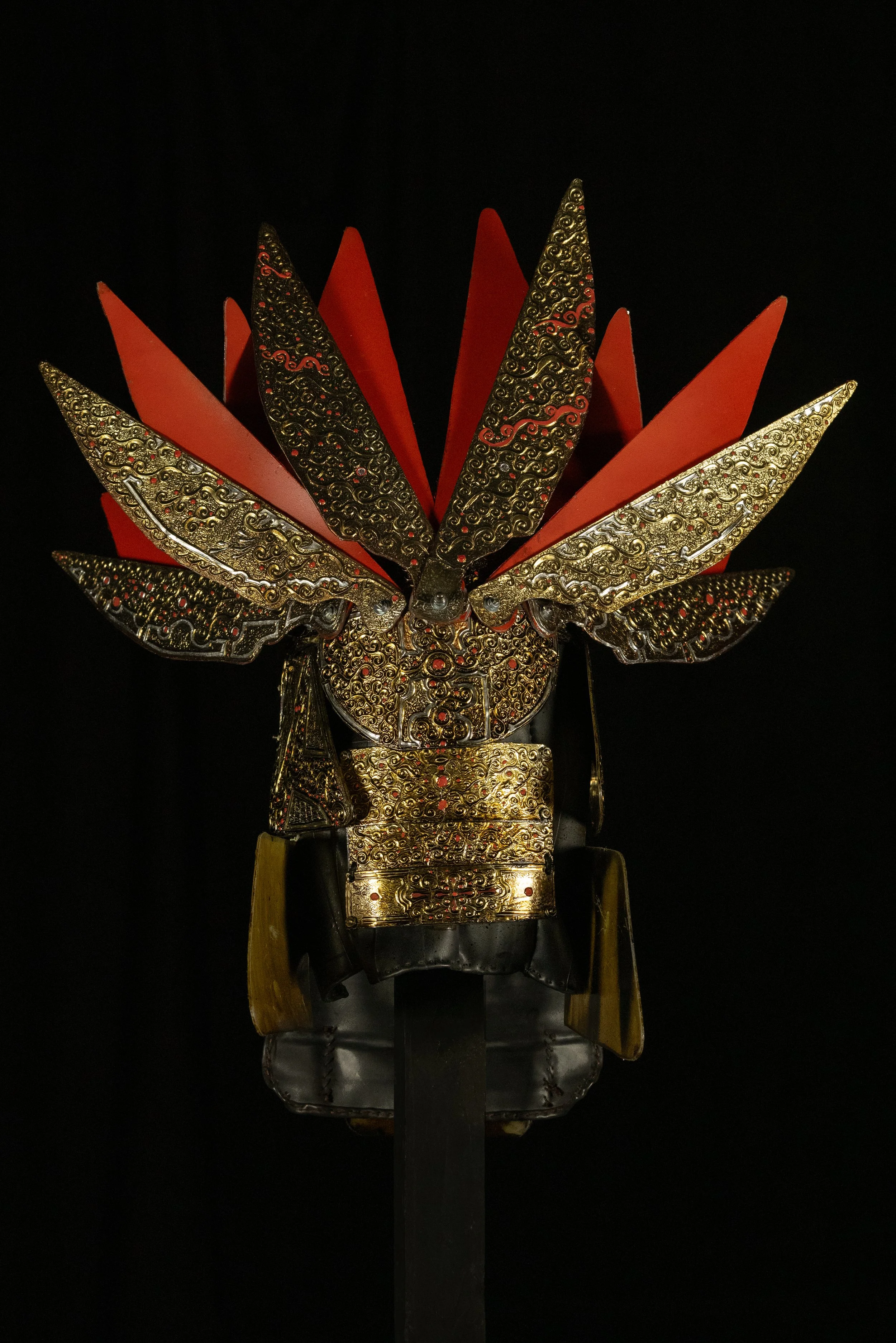 A decorative mask with intricate gold and black patterns, red accents, and wing-like protrusions against a black background.