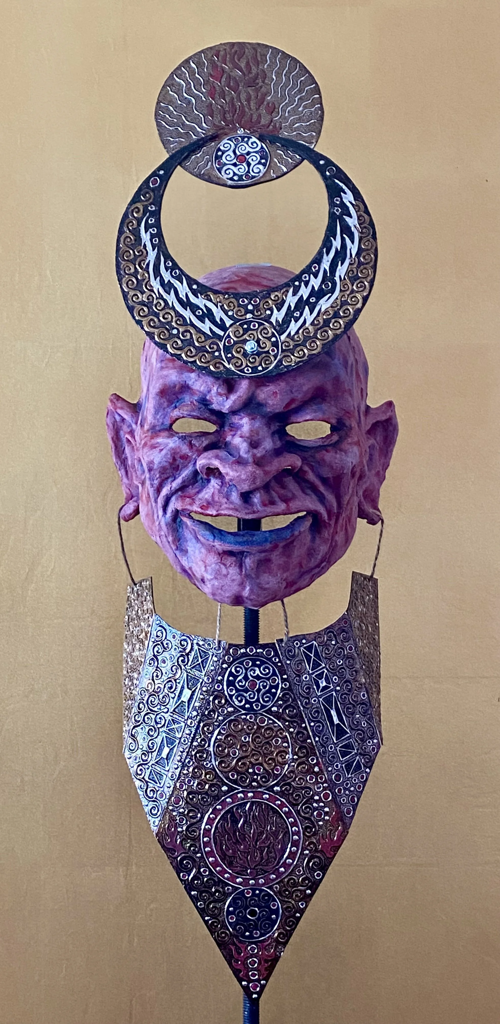 A sculpture featuring a pinkish, face-like mask with exaggerated features and an open mouth. The sculpture is adorned with ornate, circular metallic elements and intricate patterns, with the metallic pieces forming a circular headpiece and decorative