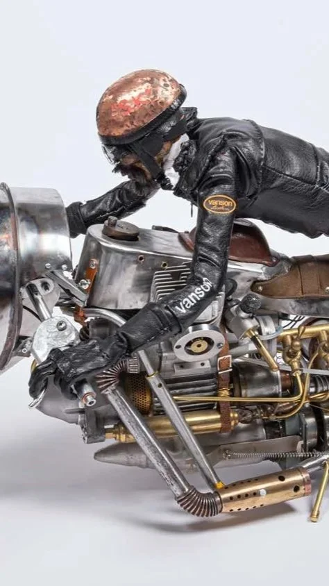 Model of a motorcyclist wearing a black leather jacket, racing helmet, and gloves, riding a vintage-style motorcycle with metallic and gold parts.