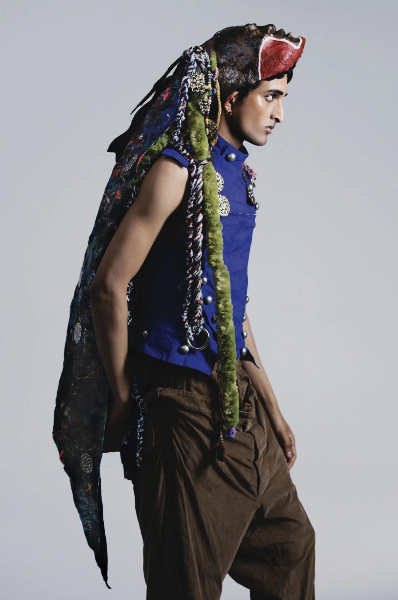 A person with dark hair and medium skin tone wearing a blue sleeveless shirt, brown pants, and a colorful, textured headpiece with fabric, beads, and other adornments, standing in profile against a plain gray background.