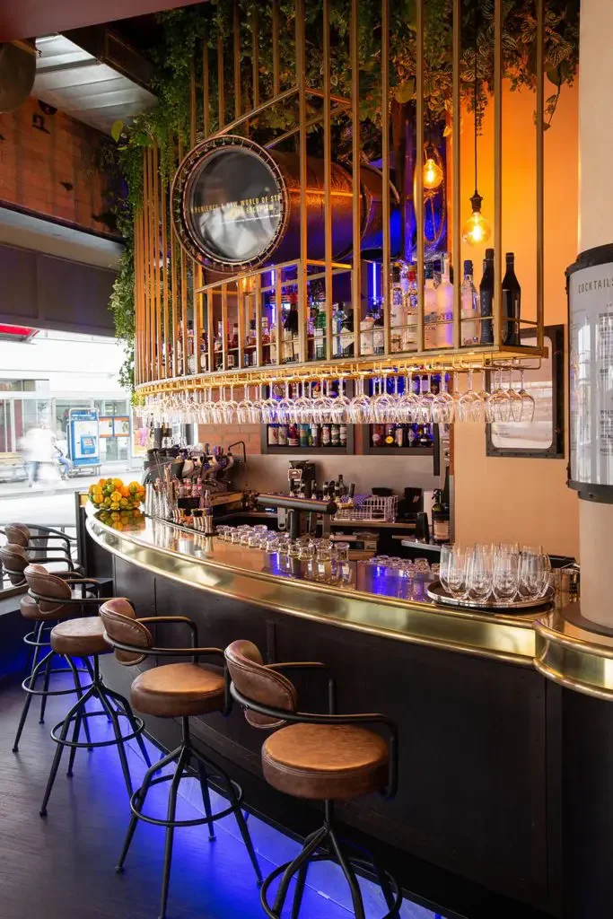 A stylish bar with a curved counter, lined with four brown leather barstools with black metal frames. Above the bar, hanging wine glasses are suspended upside down from golden racks. Shelves behind the bar display various bottles of alcohol and glass