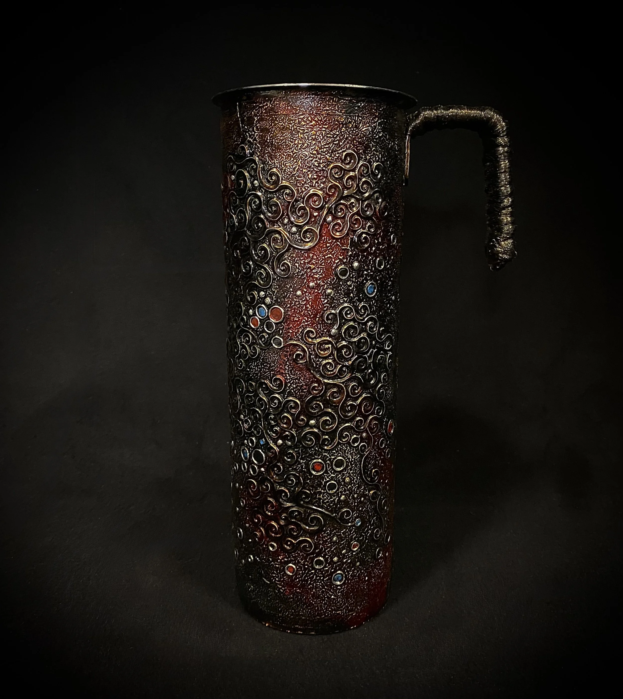 Decorative metal tank with intricate swirling design and small colored accents, set against a black background.