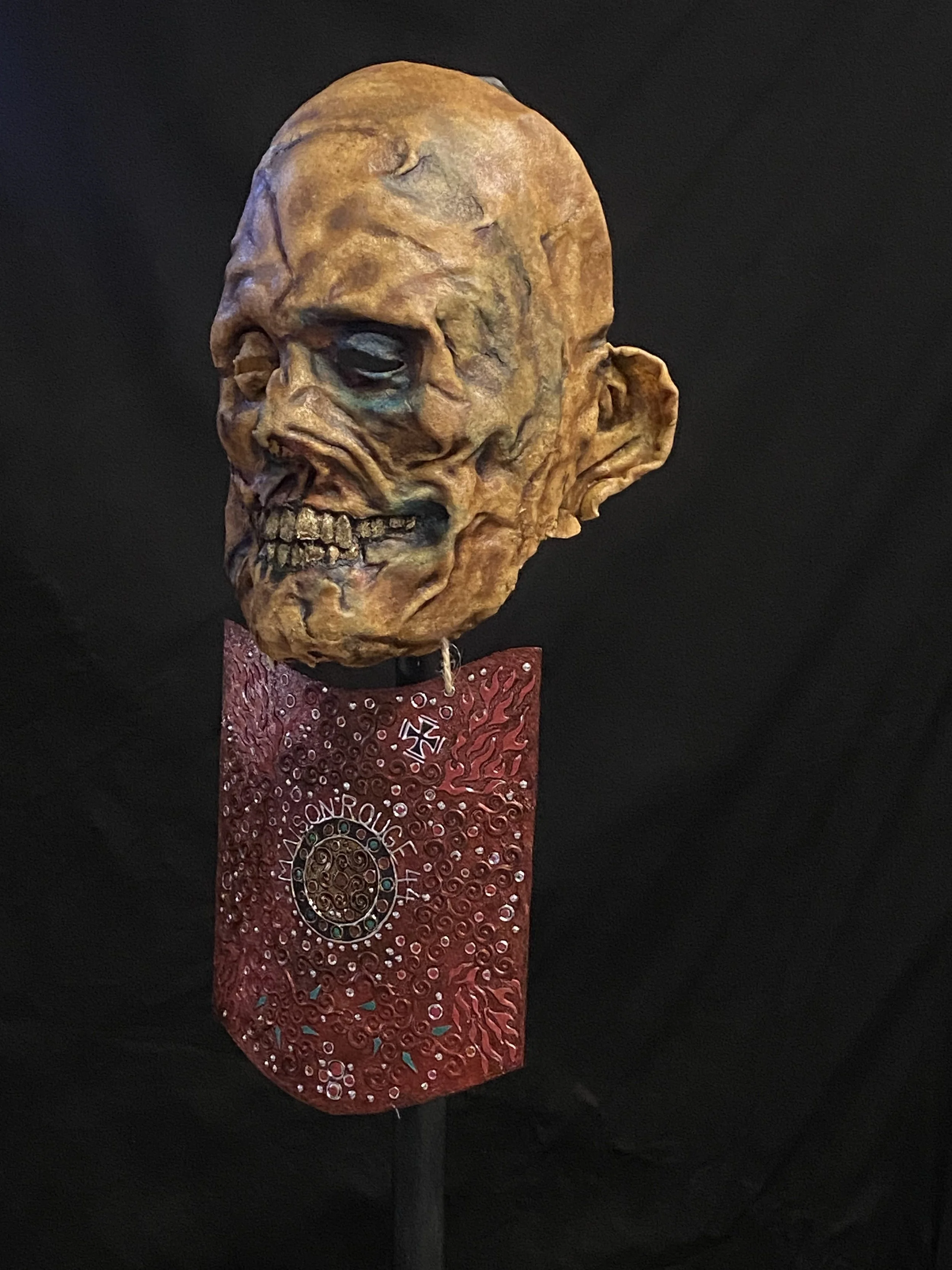 A creepy, decayed skull sculpture mounted on a stand with a red decorative plaque beneath it, set against a black background.