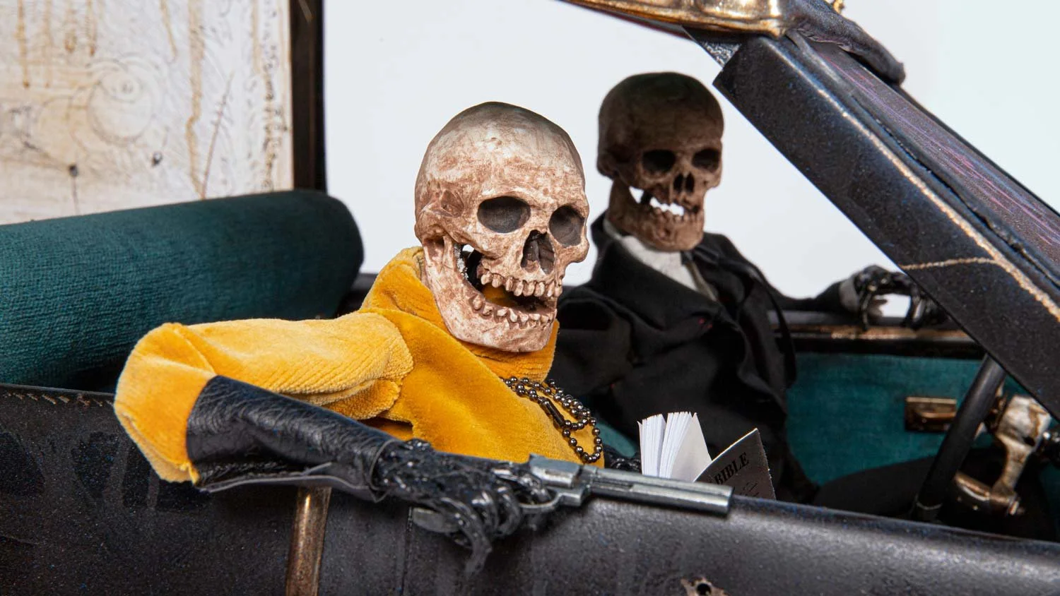 Two skeleton mannequins with skull faces seated inside a vintage car. The skeleton on the left wears a yellow jacket and a necklace, while the one on the right is dressed in dark clothing. The car's dashboard and steering wheel are visible.