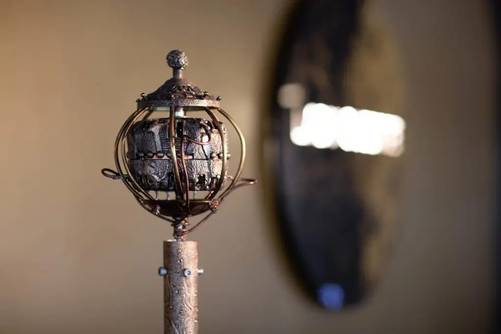 Close-up of a Victorian-style decorative scepter or wand with intricate metallic design, featuring a spherical element enclosed in a cage-like structure, standing against a blurred background.