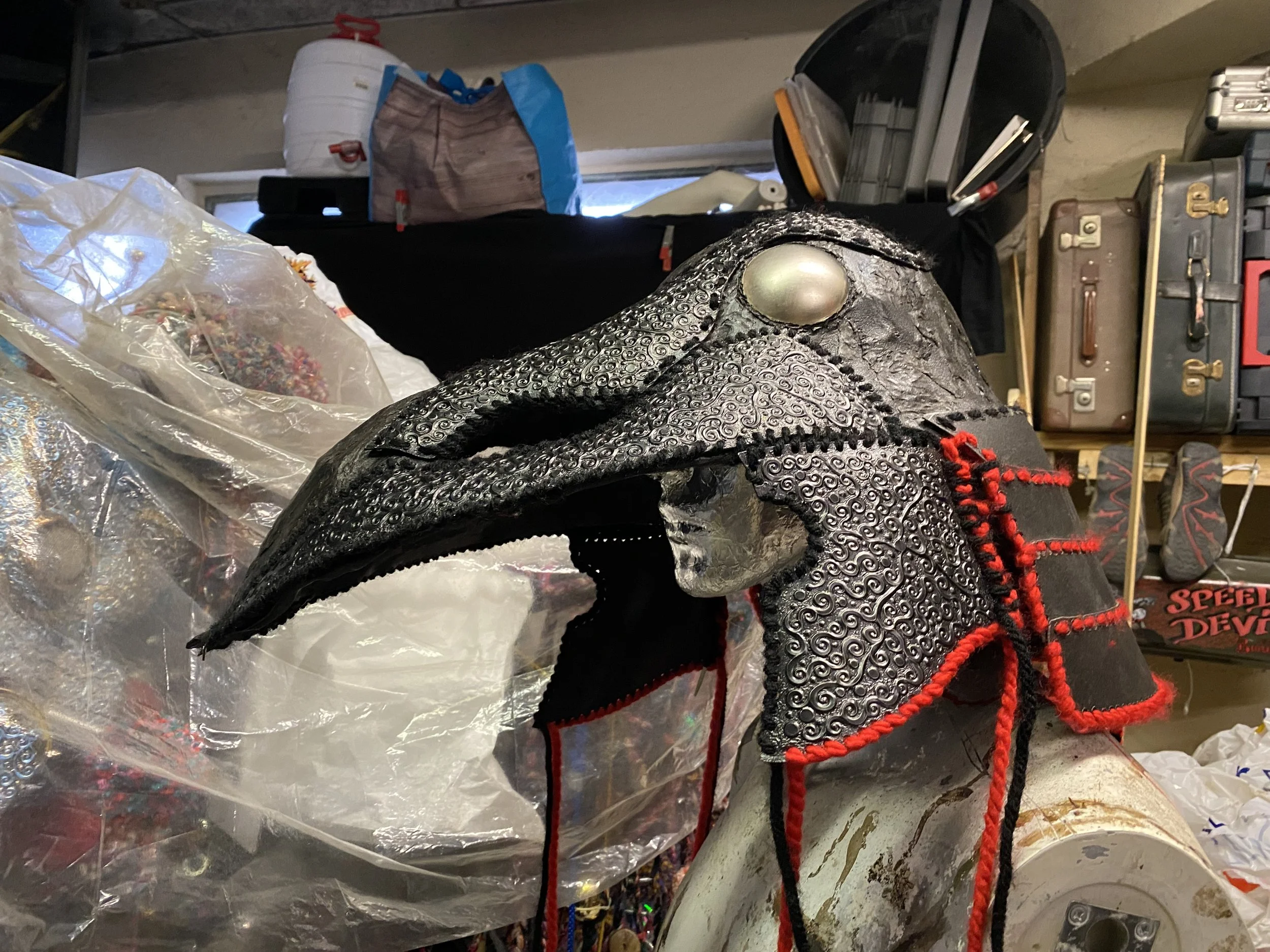 A detailed mask resembling a bird of prey, with a long beak, perched on a stand in a cluttered workshop or storage area.