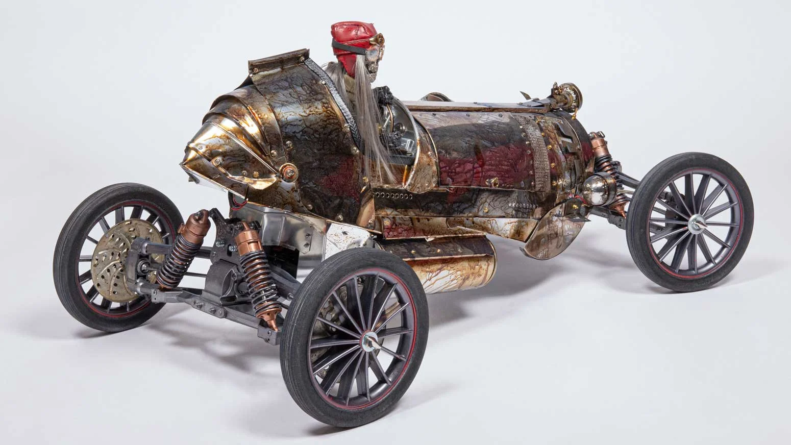 A vintage race car with a rusty, weathered metal exterior, large spoked wheels, exposed suspension, and a small driver’s seat, displayed against a plain white background.
