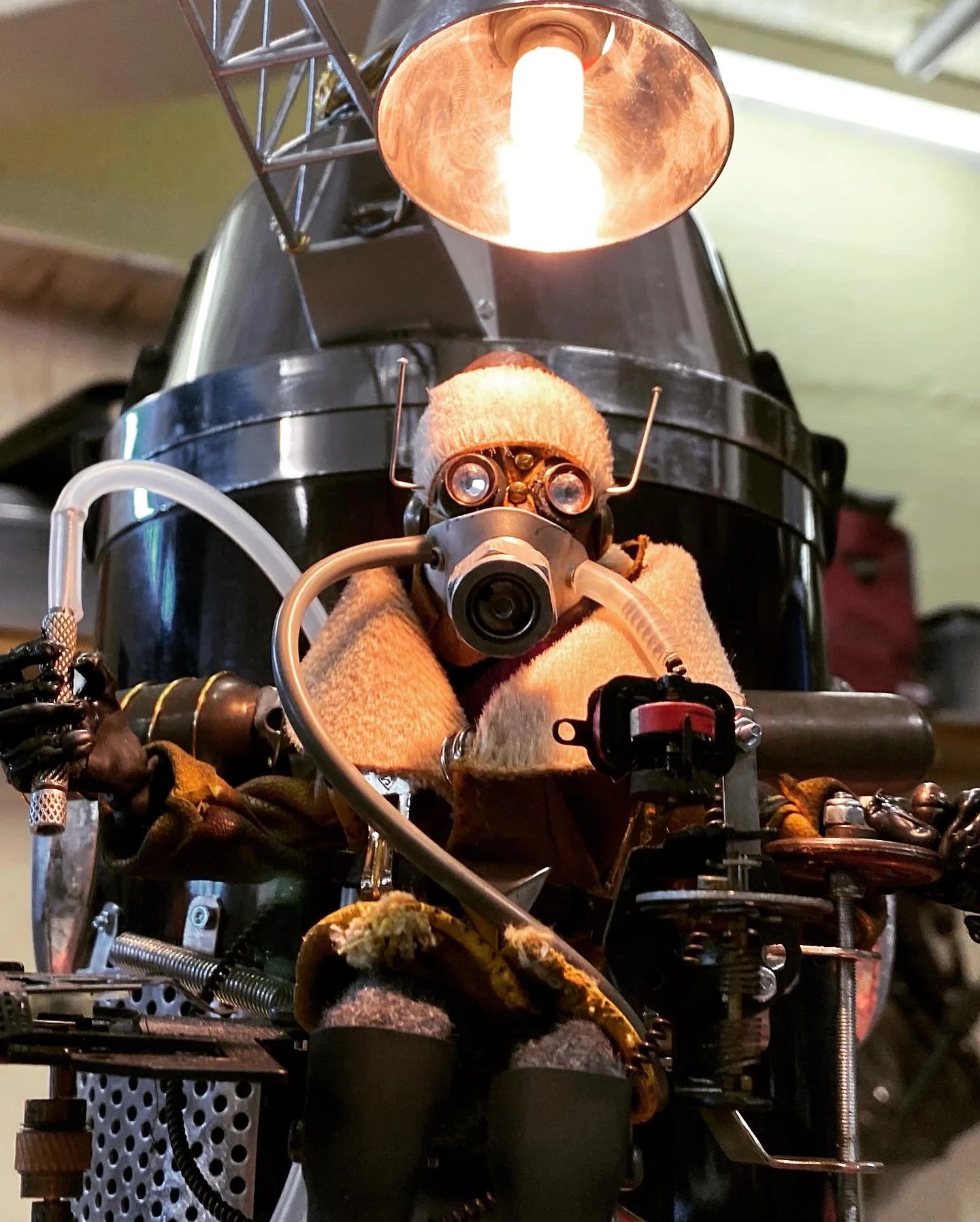 A robot with a light bulb for a head, glasses, and a face mask, dressed in clothes and sitting on a small vehicle or robot chassis, with a light shining down from above.