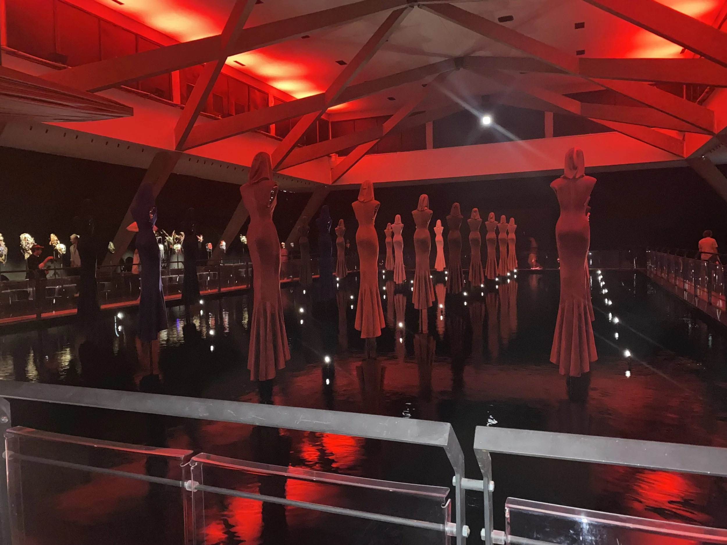 Art installation featuring tall, mannequins on long dresses on stilts reflected in water, illuminated by red lighting in an indoor space with modern architecture.