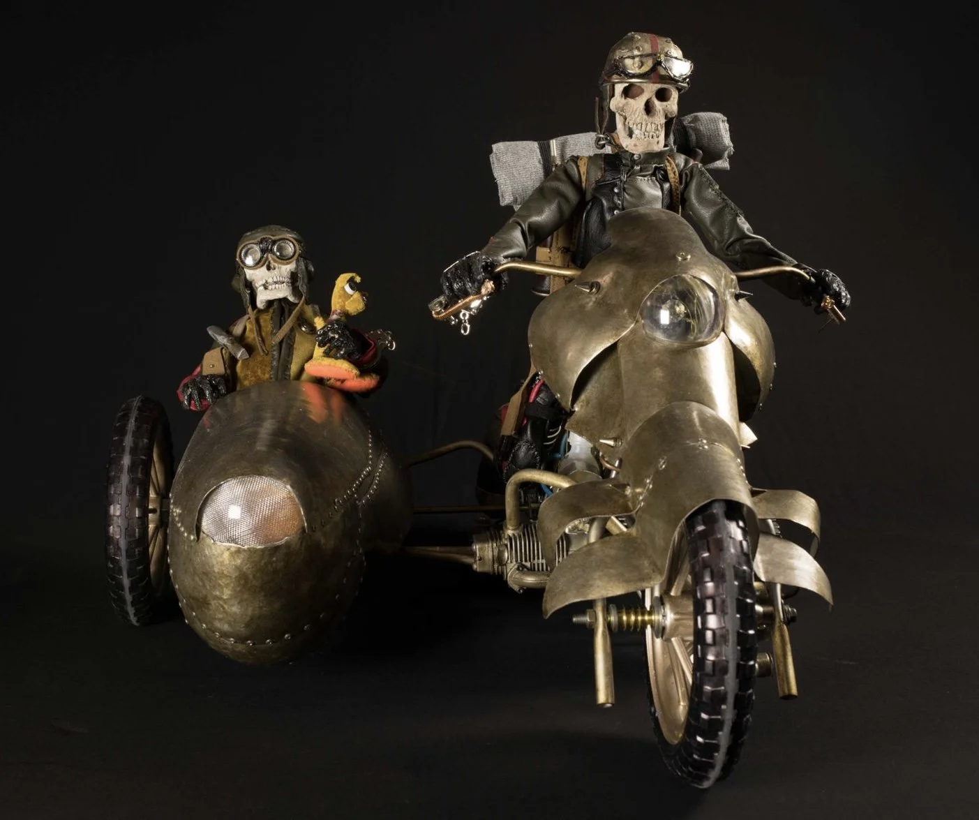 Decorative sculpture of two skeletons riding a vintage motorcycle with a sidecar, against a black background.