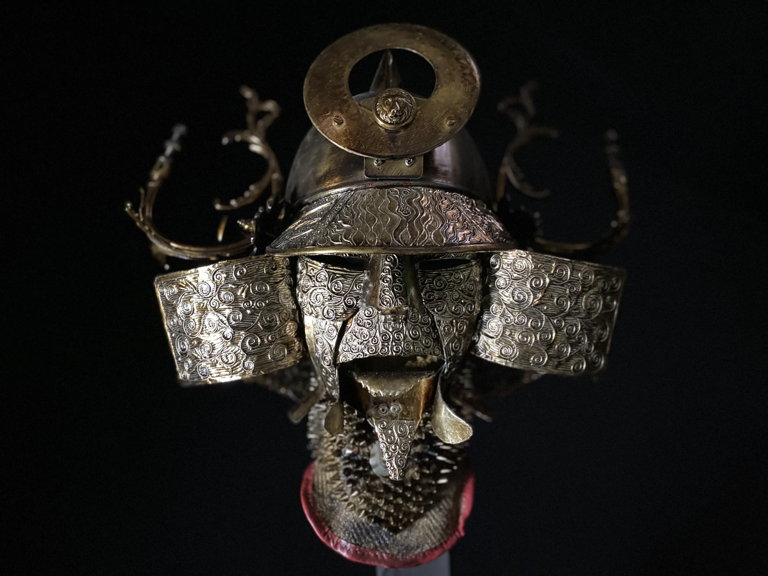 A metal sculpture of a face with intricate swirling patterns, a helmet-like headpiece, and decorative elements, set against a dark background.