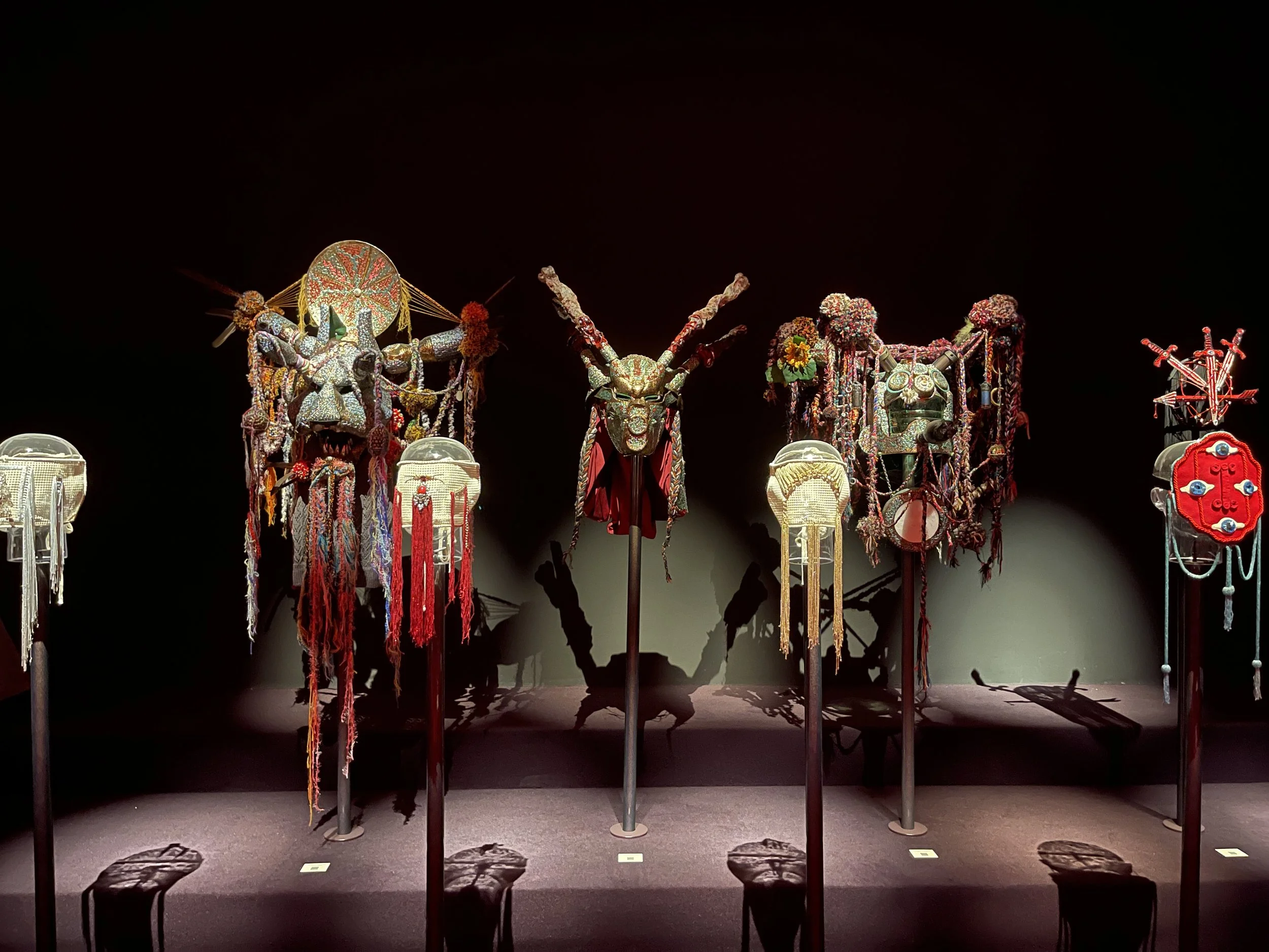 An art exhibit featuring four elaborate, colorful masks or headdresses on stands, decorated with beads, flowers, and various adornments, displayed against a dark background.