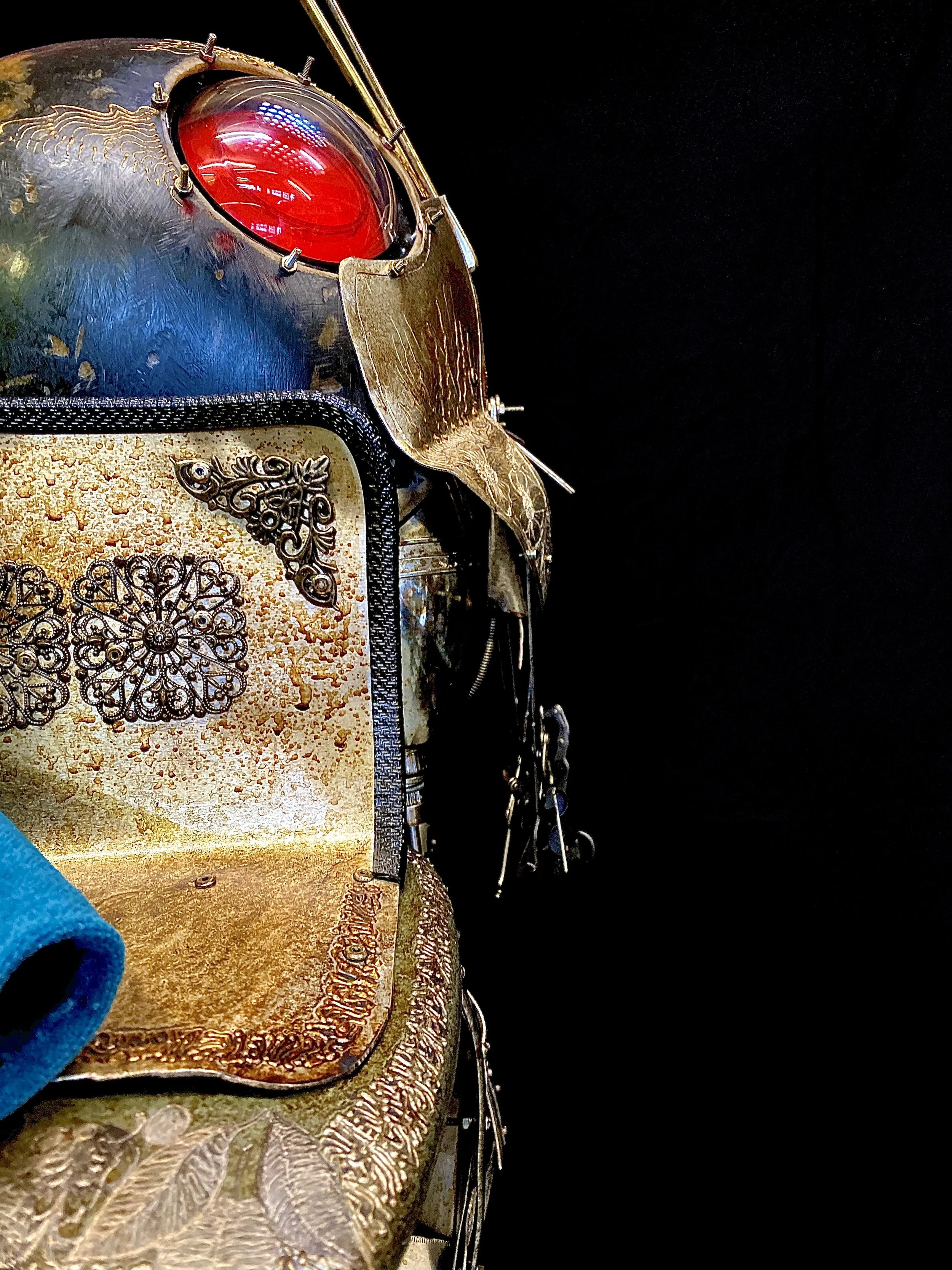 Close-up of a steampunk robot sculpture made of metal and found objects, including a large red eye, ornate patterns, and metallic details against a black background.