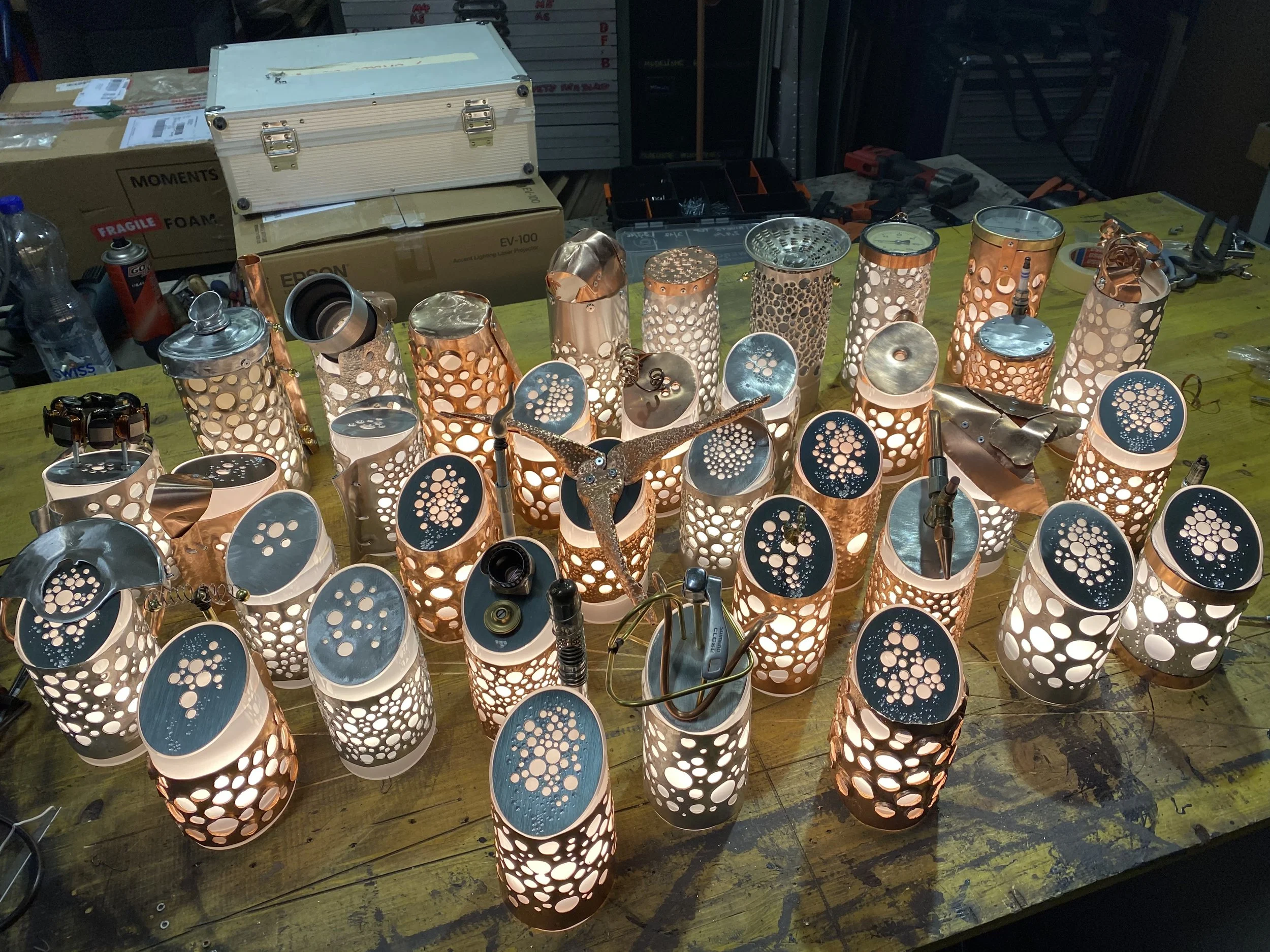 A collection of decorative table lamps with perforated metal covers emitting warm light, arranged on a workshop table with tools and boxes in the background.