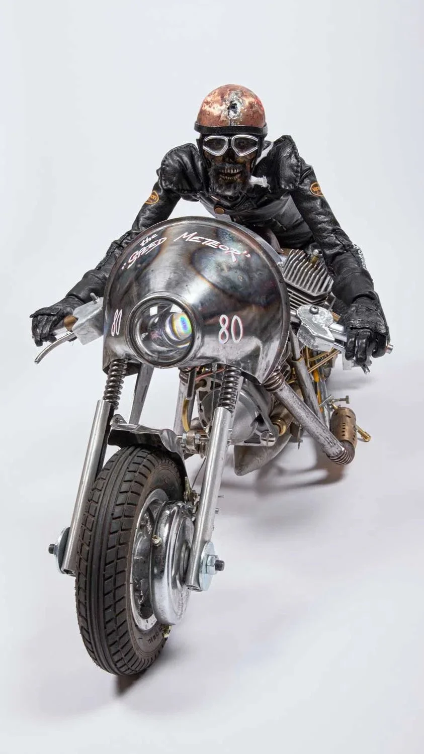 A person in a black leather jacket and gloves, wearing a rusty helmet and goggles, riding a metal motorcycle with a large front headlight and minimalist design.