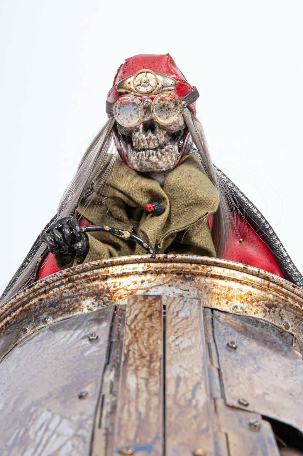 A humanoid skeleton wearing a red helmet with goggles, a khaki scarf, and a red jacket, riding a motorcycle from a low-angle perspective.