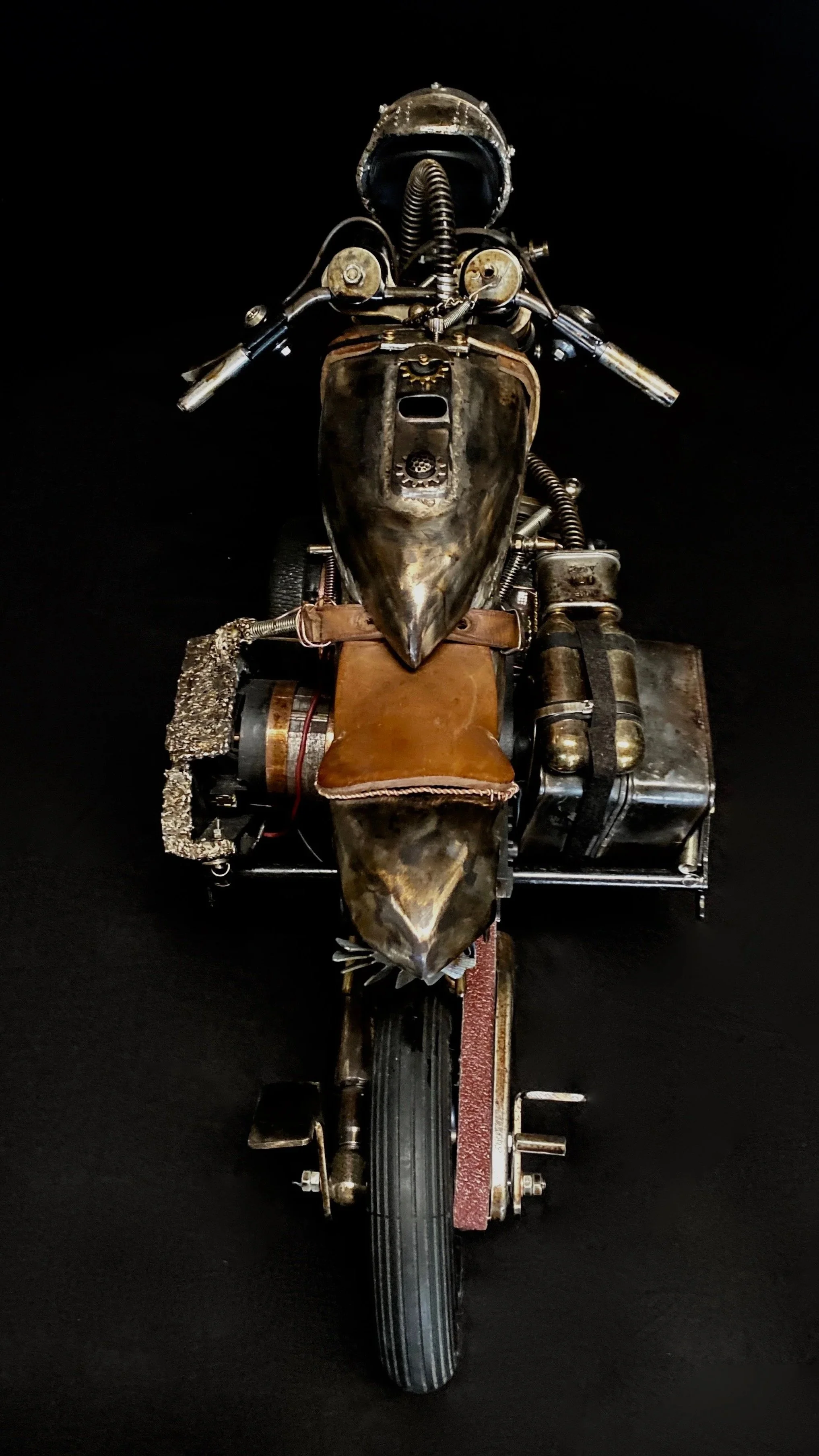 Front view of a motorcycle constructed with a steampunk aesthetic, featuring metallic parts, leather accents, and vintage-style components, set against a black background.