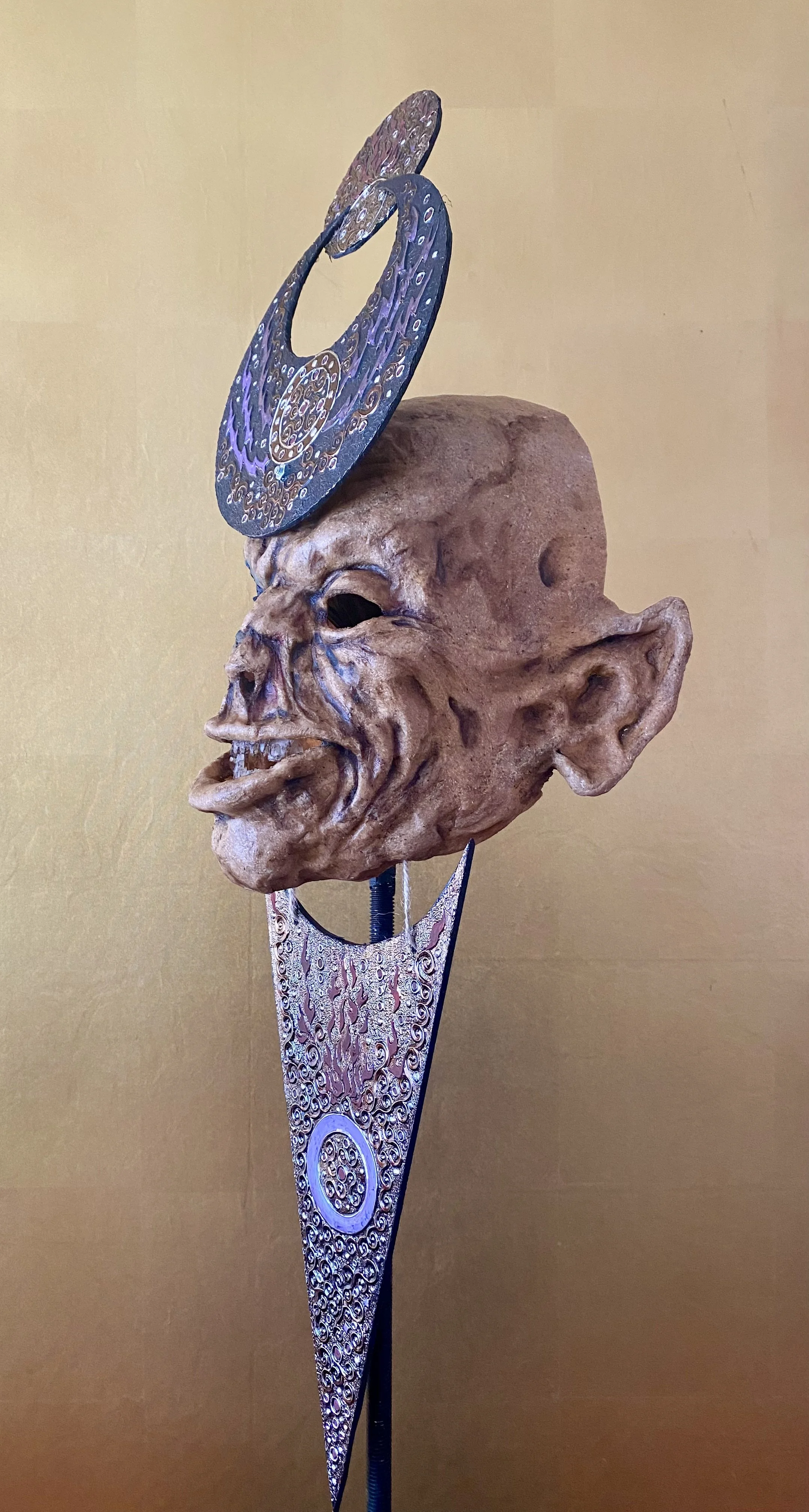 An art sculpture featuring a grotesque, exaggerated human face with large ears and an open mouth, mounted on a stand with decorative elements. It has a metallic spiral ornament above its head.