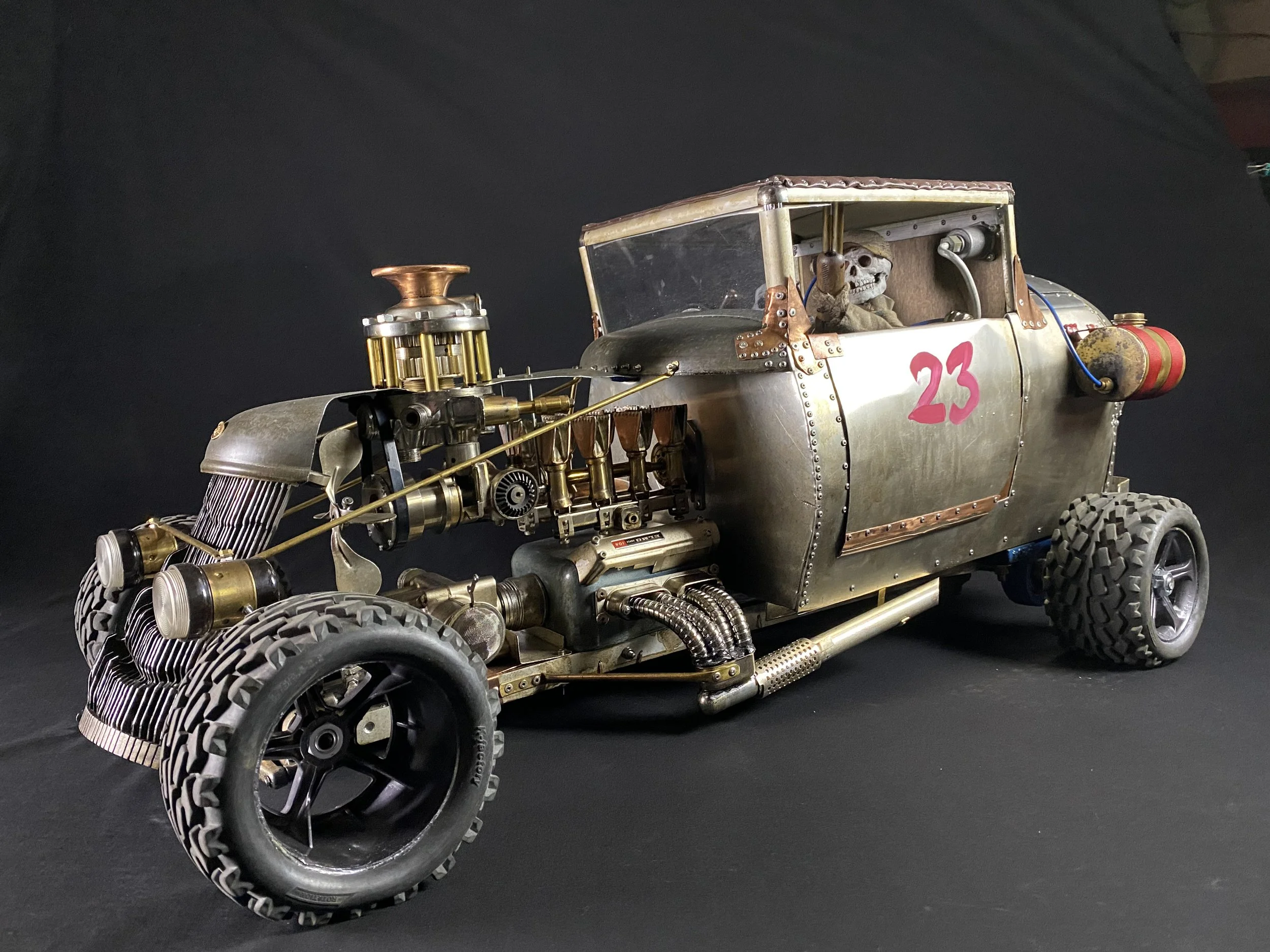 A vintage, custom-built hot rod car with a skeleton driver wearing goggles, featuring exposed mechanical parts, brass and metal accents, and the number 23 painted on the side.