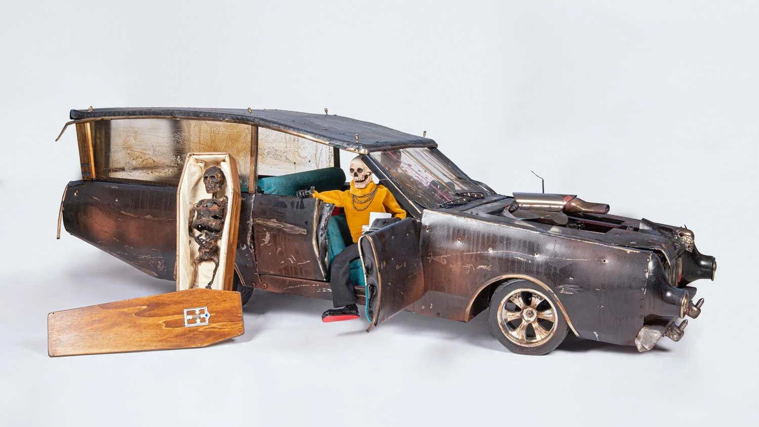 A sculpture of a damaged car with skeleton figures inside. One skeleton in a yellow shirt is sitting in the driver's seat, and another skeleton is standing in the back near the open door, with the lower half of the skeleton visible. The car is rusty 