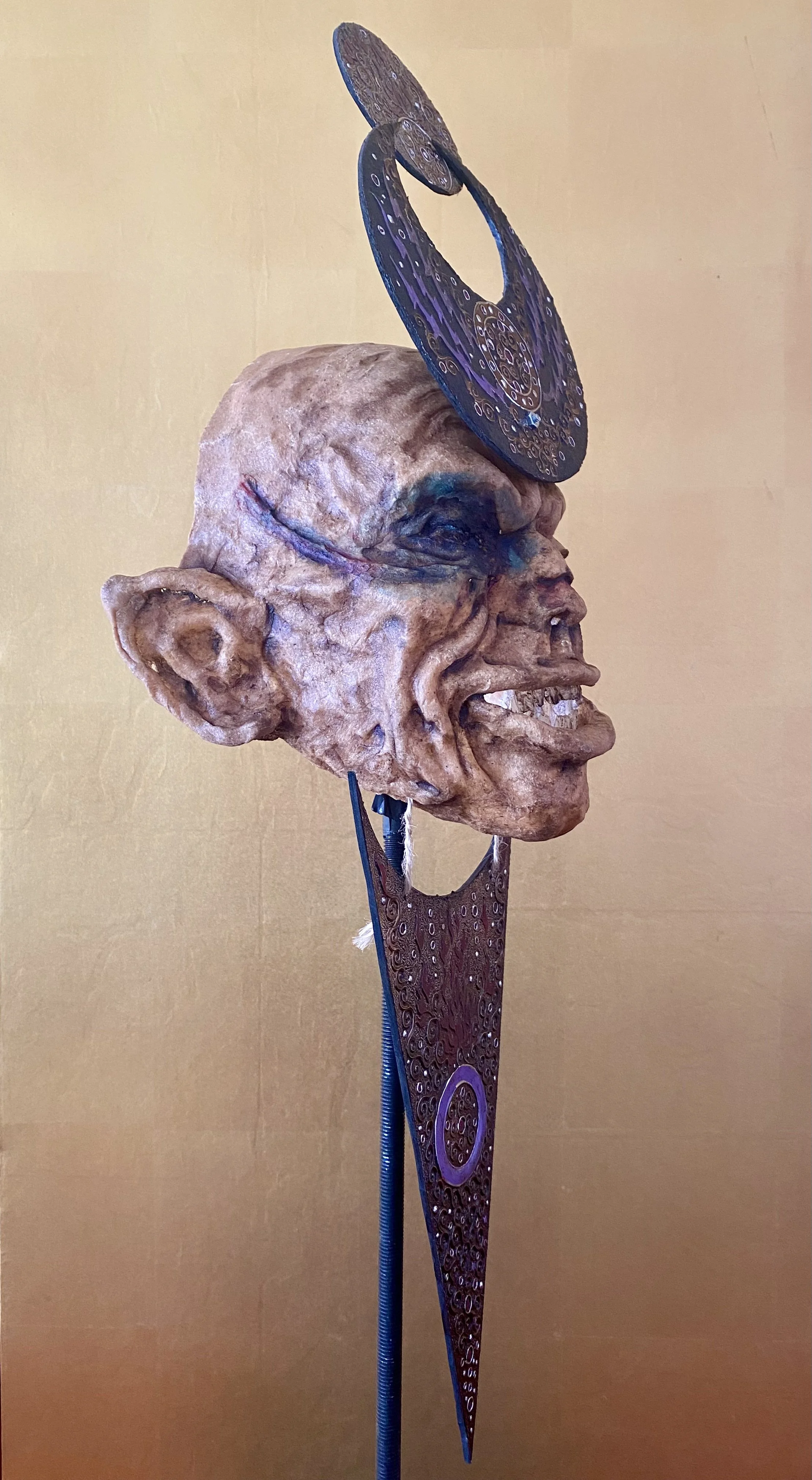 An artistic sculpture featuring a human head with exaggerated features, a large ear, and blue face paint, mounted on a stand with a circular and oval-shaped decorative element above the head.