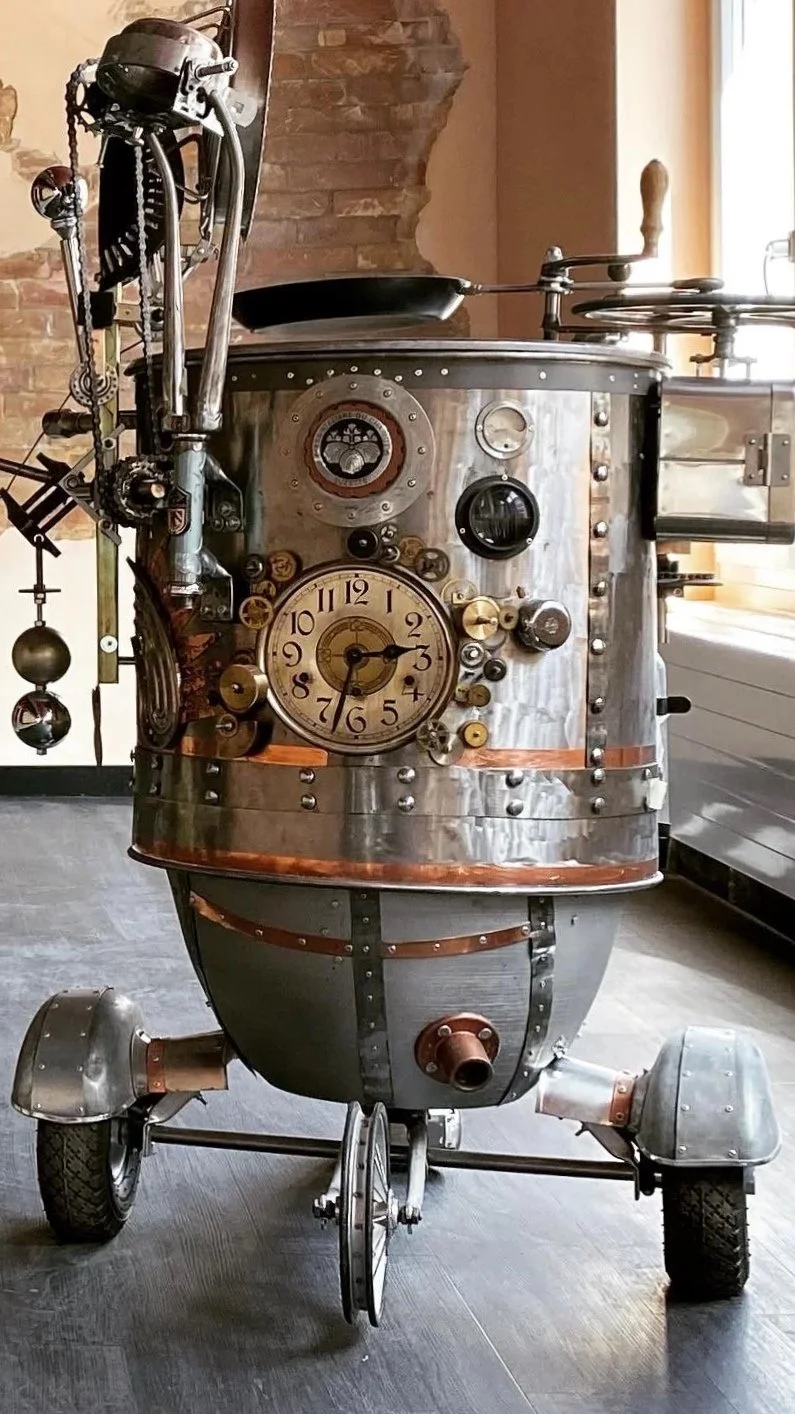A robot made from various metallic parts, featuring a clock on its chest, small wheels, and mechanical components, set indoors near a window and brick wall.