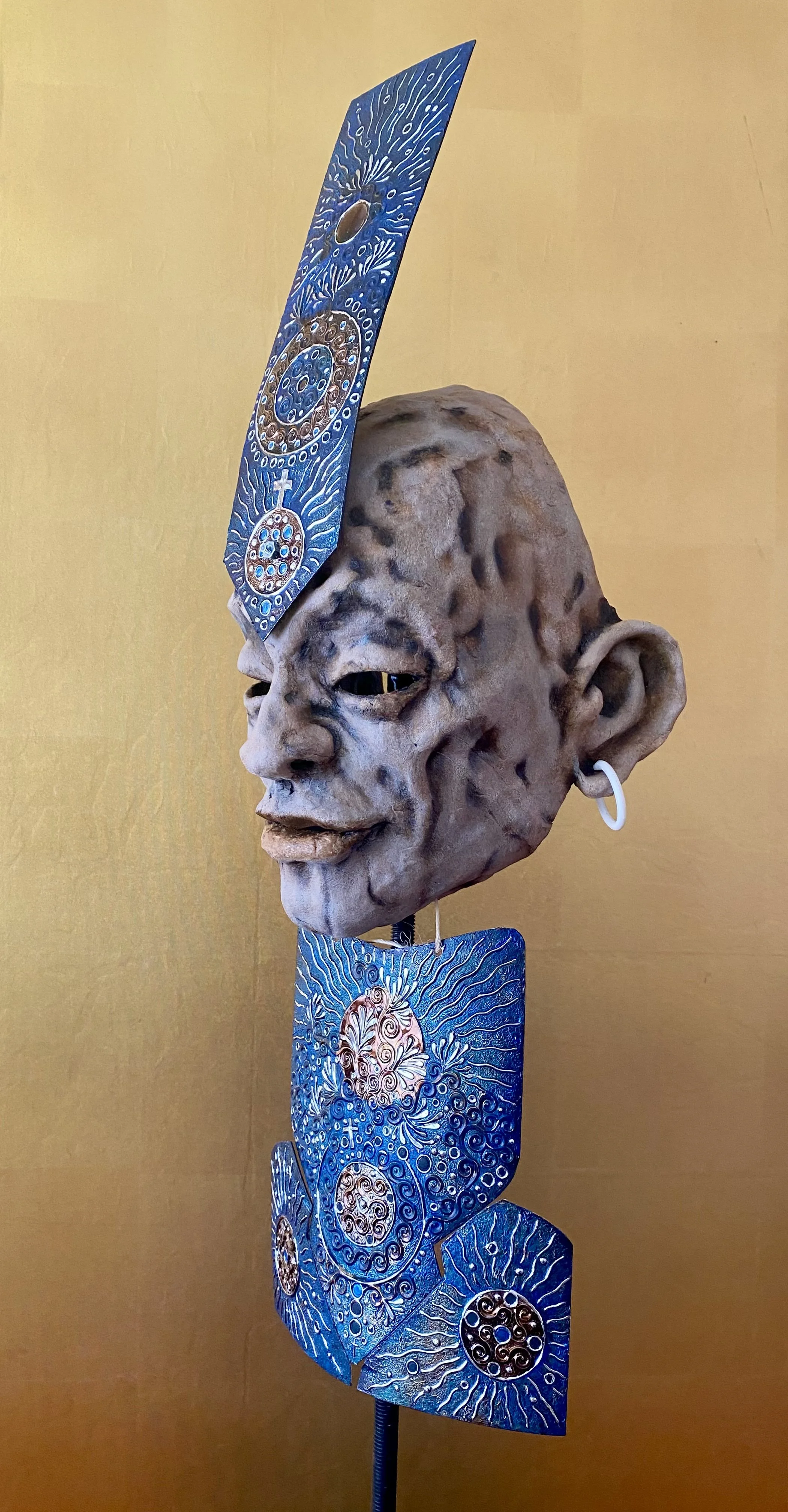 An art sculpture of a human face with exaggerated features, including large ears and textured skin, wearing artistic jewelry and headpieces, against a yellow background.