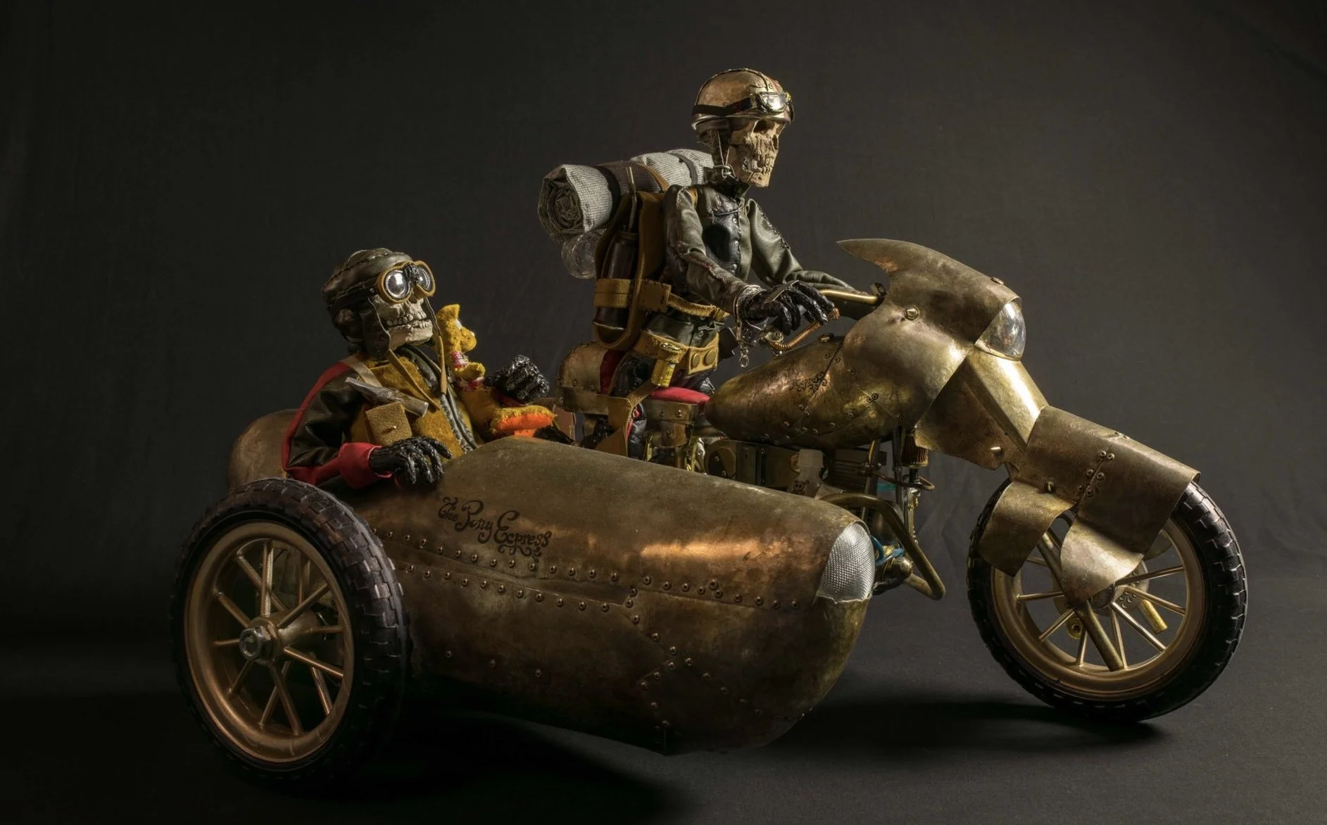 A detailed sculpture of a skeleton riding a vintage motorcycle with a sidecar, both made of metallic materials, against a black background.