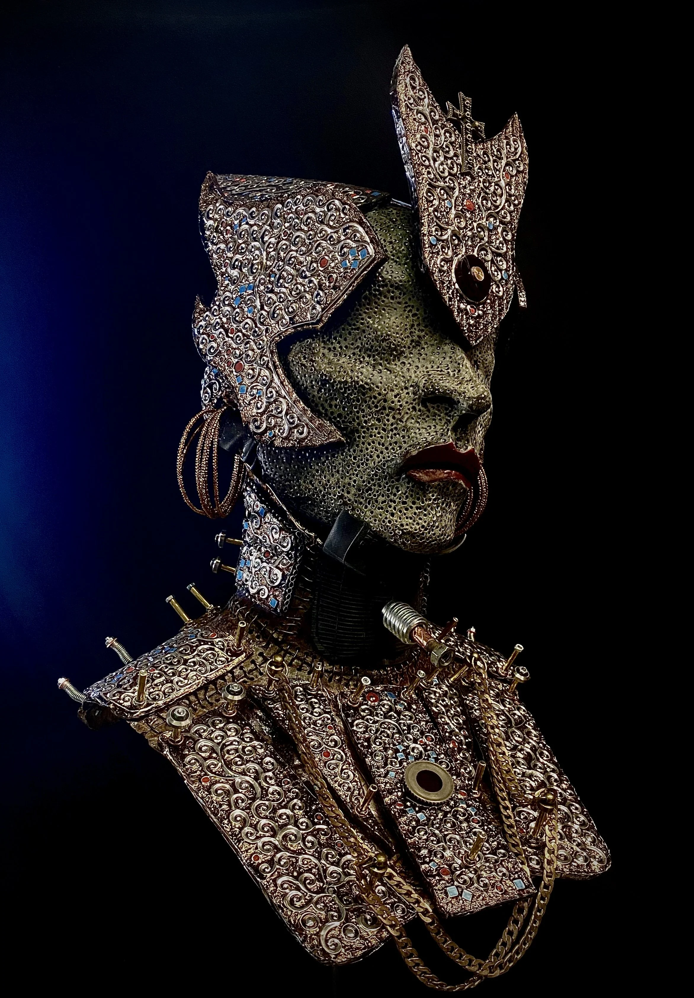 A futuristic sculpture of a woman's bust with metallic and jeweled adornments, featuring a textured green face, intricate silver and gold embellishments, and a dark background.