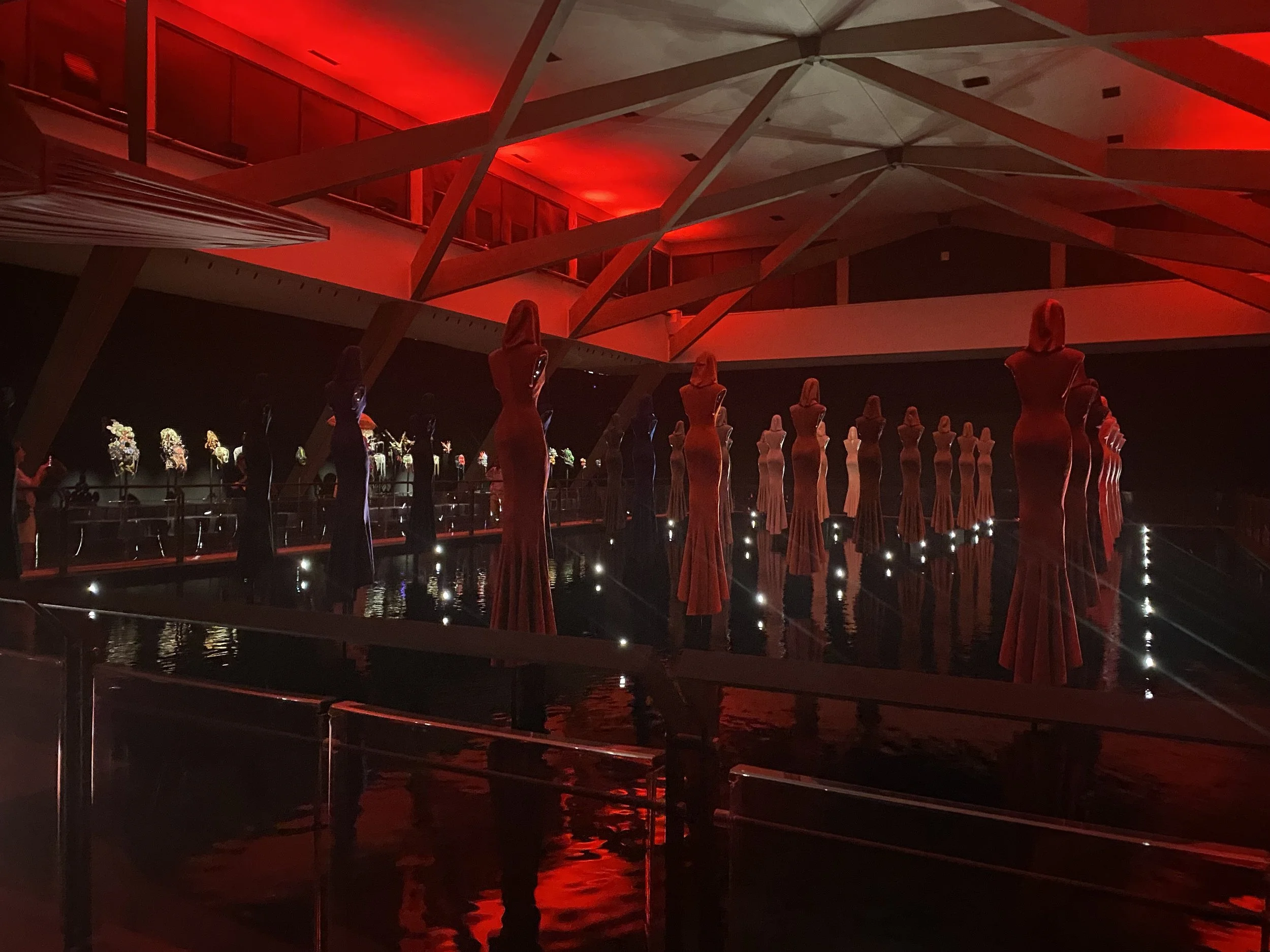 Indoor art installation featuring mannequin-like figures in long dresses, illuminated with red lighting, surrounded by water with small lights, and decorated with floral arrangements in the background.