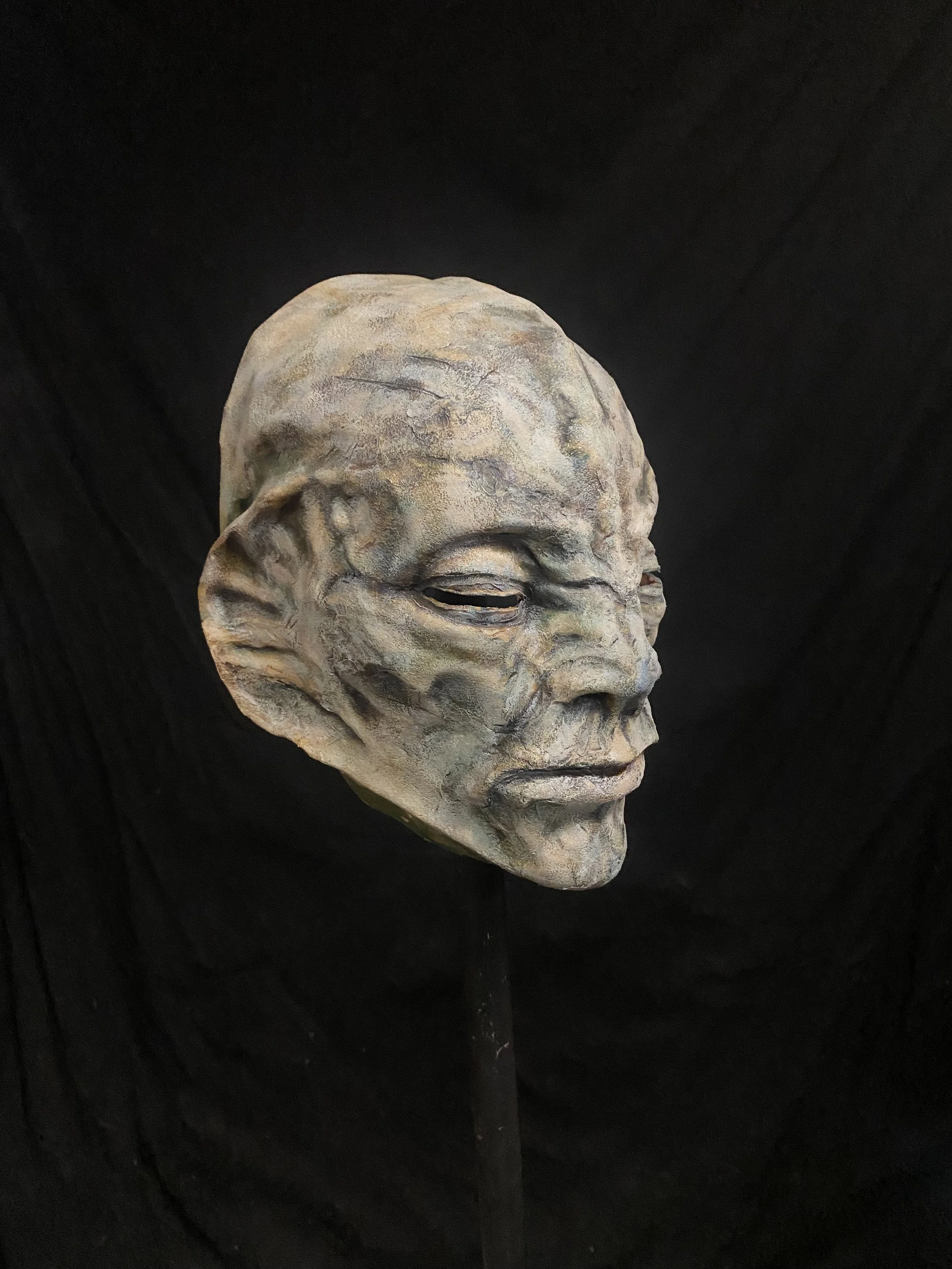 A sculpture of an alien or fantasy creature's head with textured skin, elongated ears, and closed eyes, displayed against a black background.